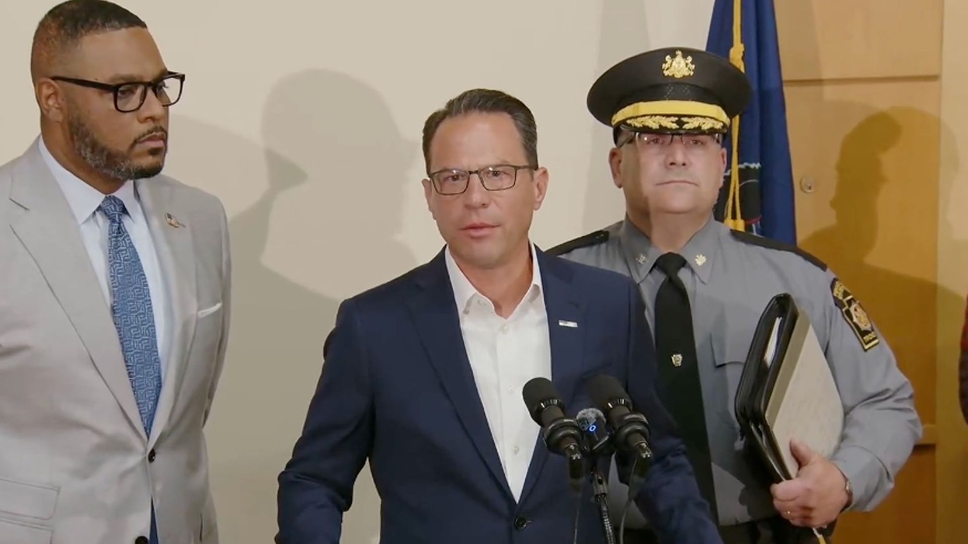 Three men at a press conference; the center man in a navy suit speaks at a podium with Pennsylvania emblem, flanked by a man in a light gray suit and a police officer in uniform holding a folder.