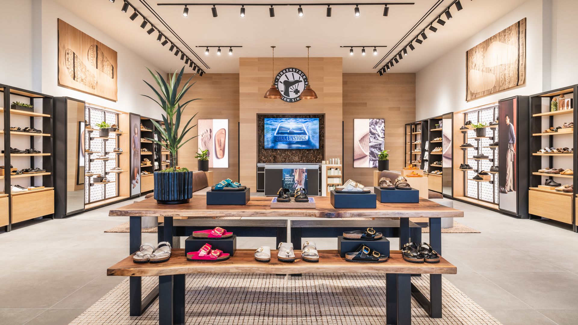 Interior of a modern shoe store with wooden shelves, sandals on display, a central wooden wall featuring a circular Birkenstock logo and screen, copper pendant lights, and green plants.