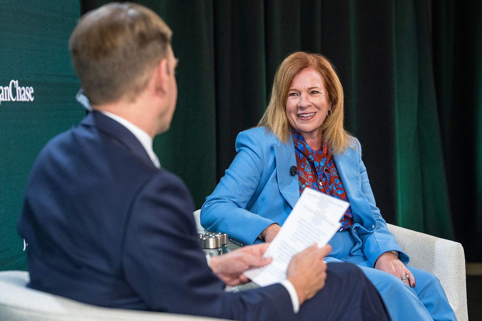 Panel discussion: a smiling blonde woman in a light blue suit and red patterned blouse chats with a man in a dark suit; he holds papers as she leans forward on a green stage backdrop.