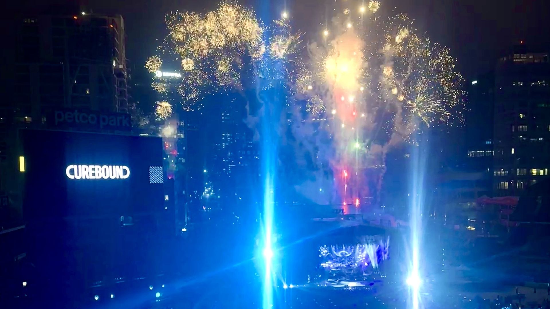 A firework display over an elton john concert at Petco Park, with downtown San Diego behind the stage and the words "curebound" on a Jumbotron