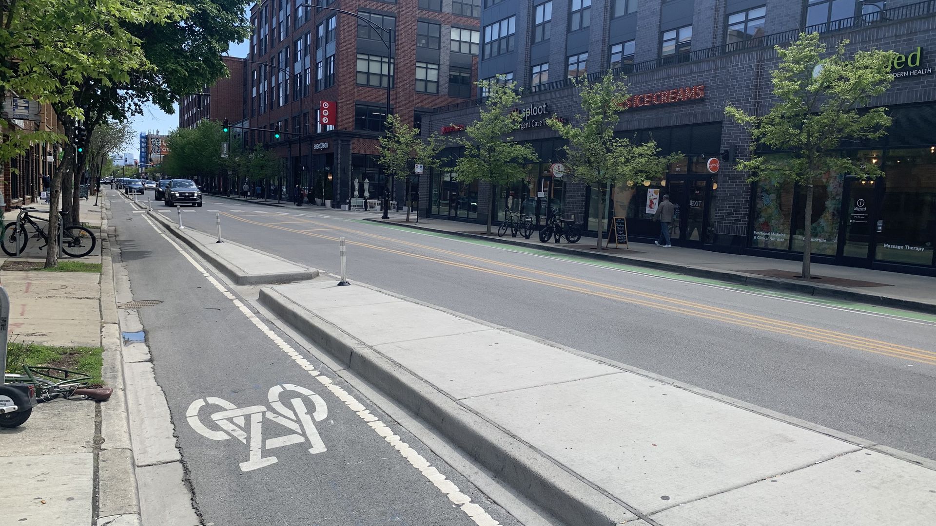 Sunny urban street with a bike lane, bicycles parked along the sidewalk, and brick and gray buildings. Trees line the street; storefronts and a few cars are on the road.