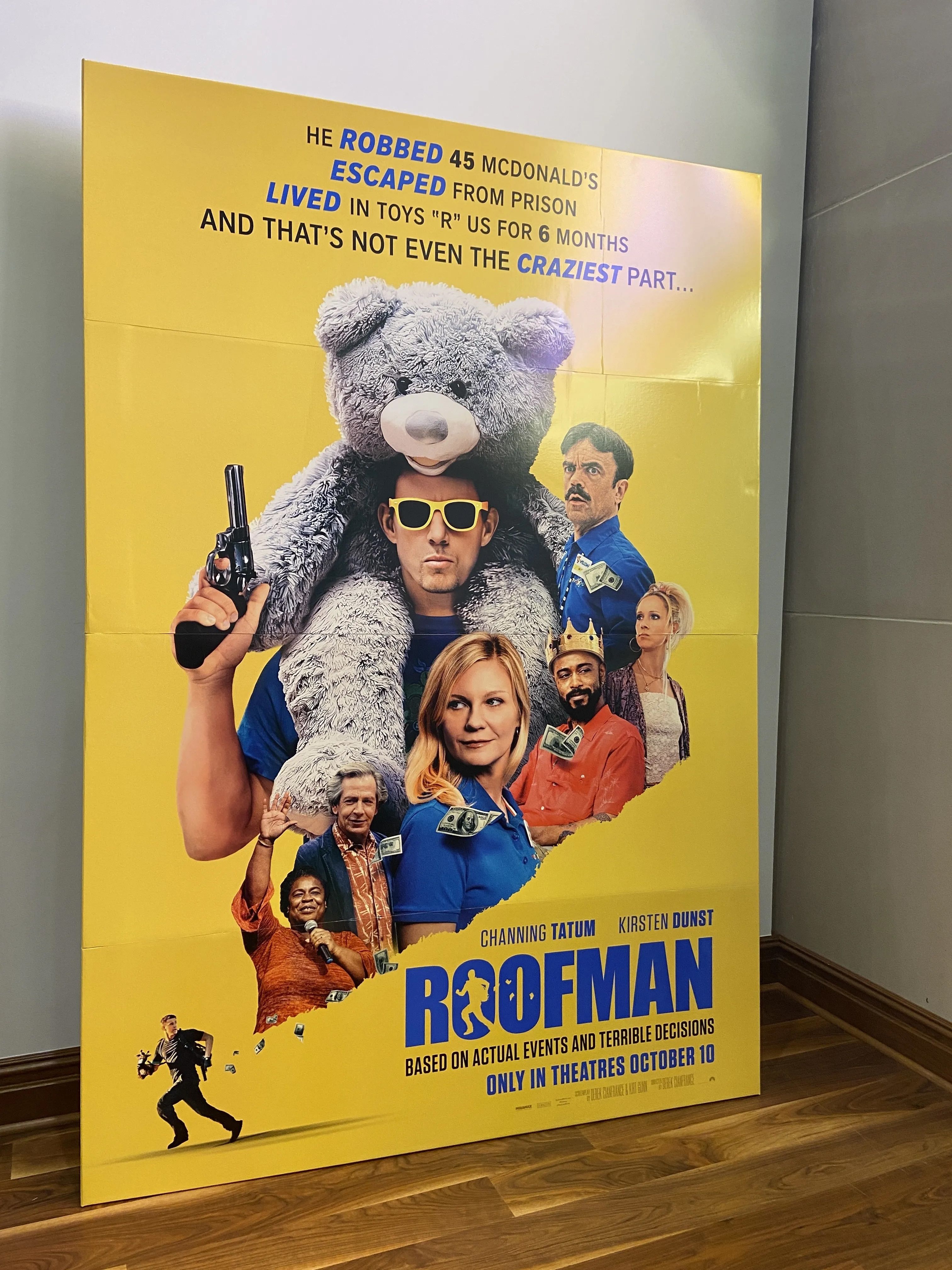 Bright yellow poster for the movie "Roofman" showing a man with a large teddy bear on his shoulders, holding a gun and wearing yellow sunglasses, surrounded by other characters with money flying around.