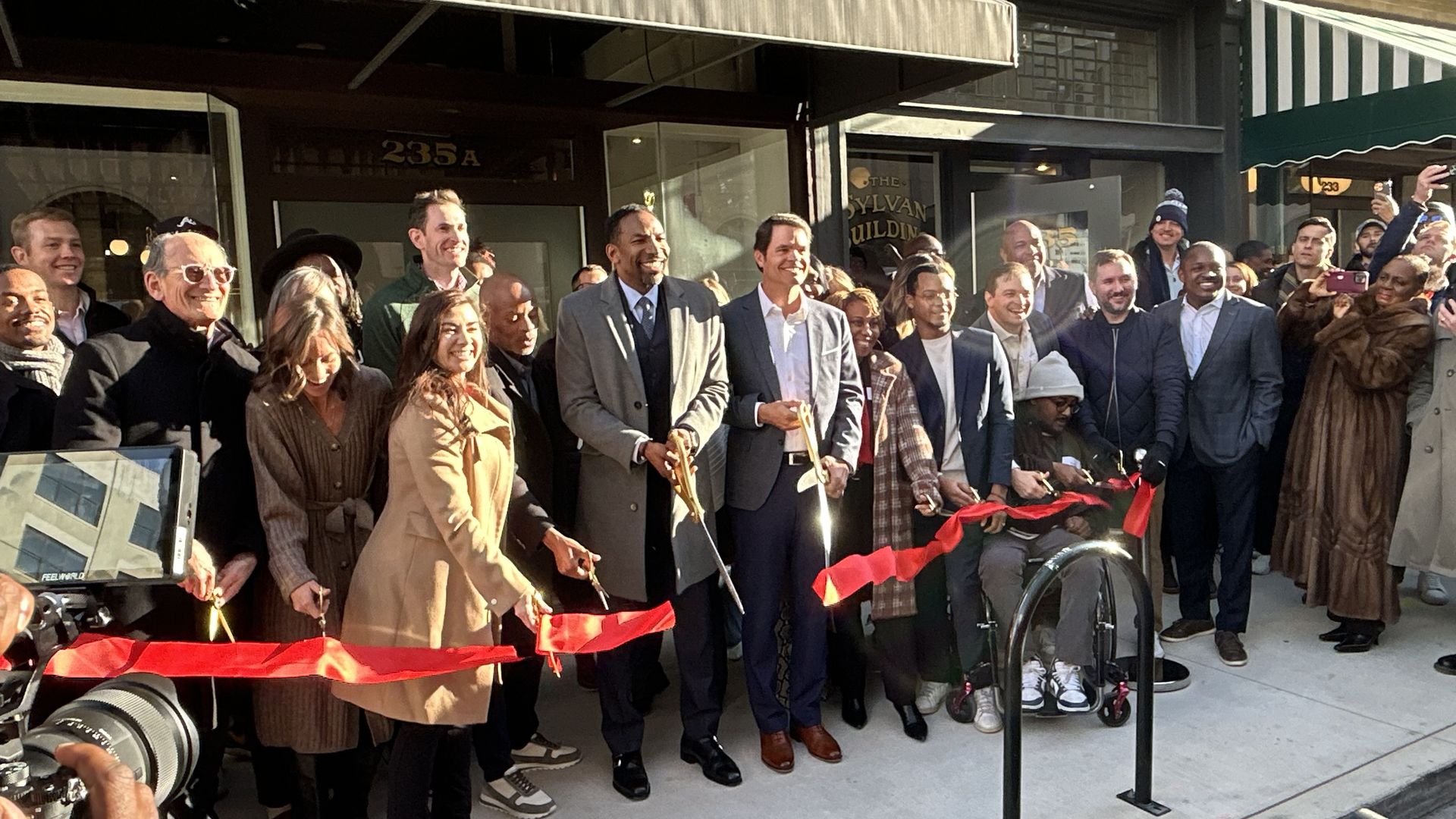 A group of people wearing winter clothing cut a large ceremonial red ribbon in front of a business incubator in a traditional urban neighborhood setting