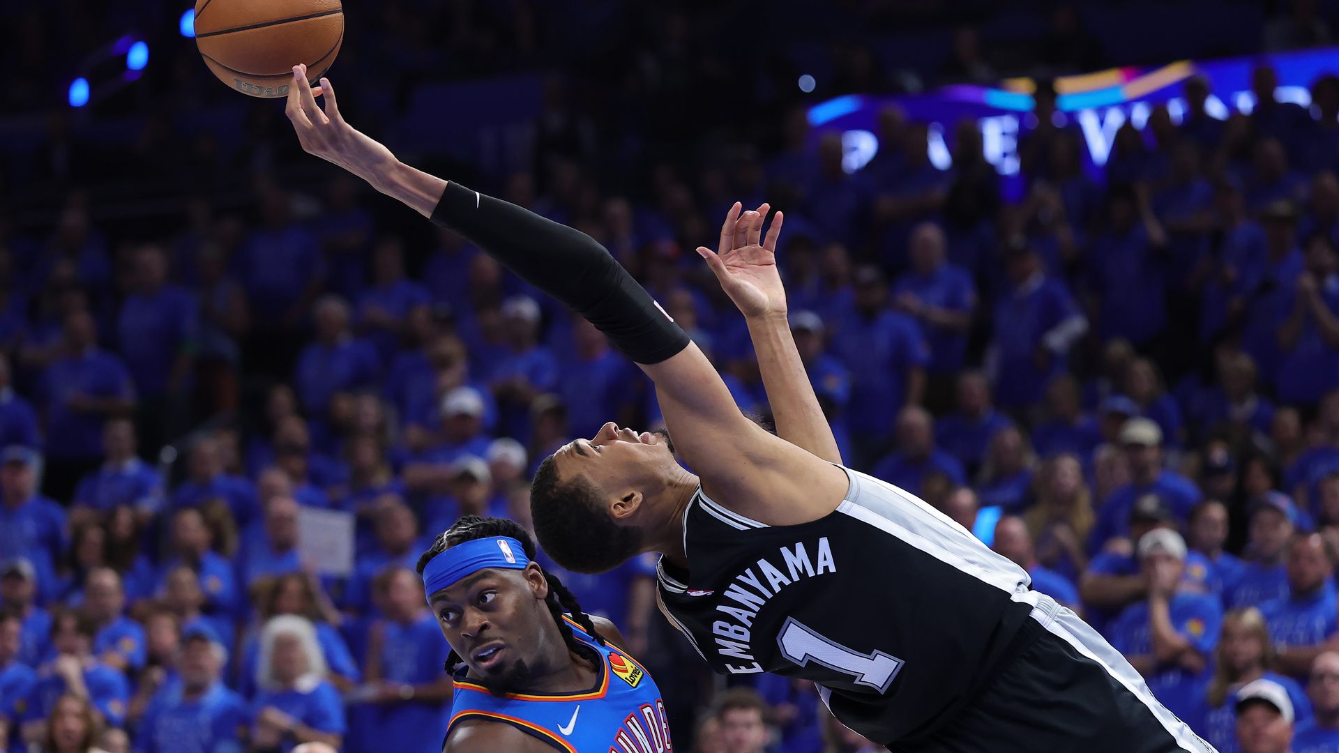 Two basketball players leap for the ball in midair during a game; one in a dark jersey reaches with an extended arm, the other in blue defends; blue-clad crowd watches in the background.