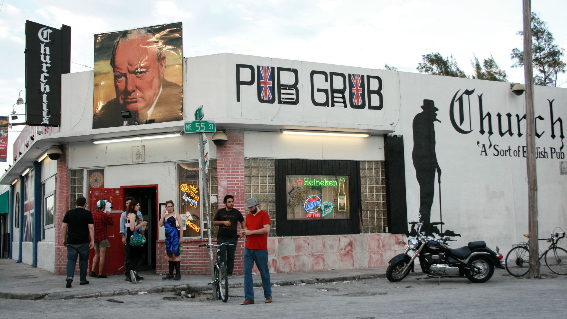 Exterior of Churchill pub, a corner building with a large painting of Winston Churchill, British flag letters spelling PUB GRUB, neon beer signs, and a black silhouette of Churchill on the wall.