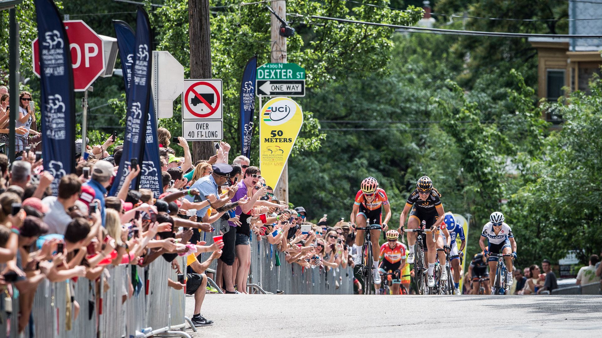 Crowd-lined street at a cycling race finish; cyclists in colorful jerseys race past cheering spectators, barriers, blue banners, a red stop sign, and a Dexter St sign with 50 meters marker.