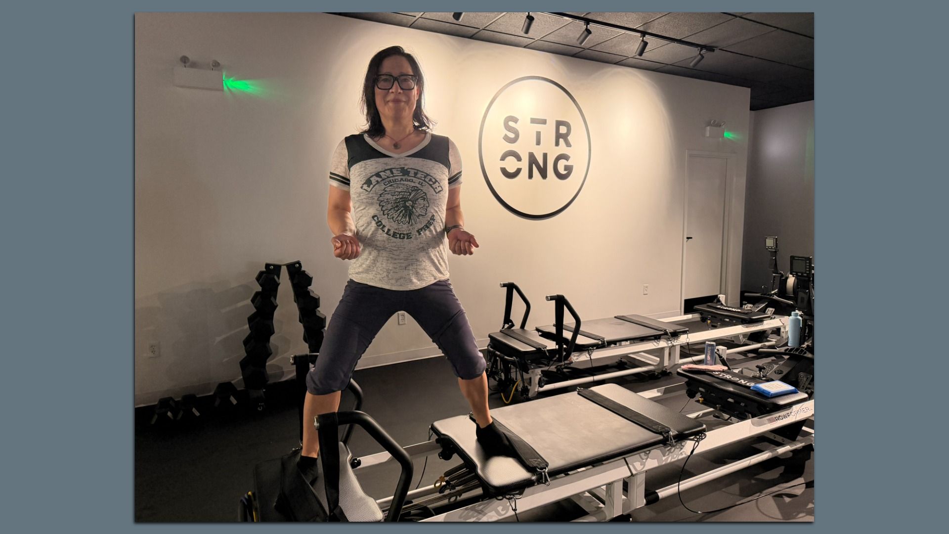 A woman with glasses stands on a Pilates reformer in a gym, wearing a gray graphic T-shirt. She grips the handles with bent elbows, in front of a large circular "STRONG" logo on the white wall.