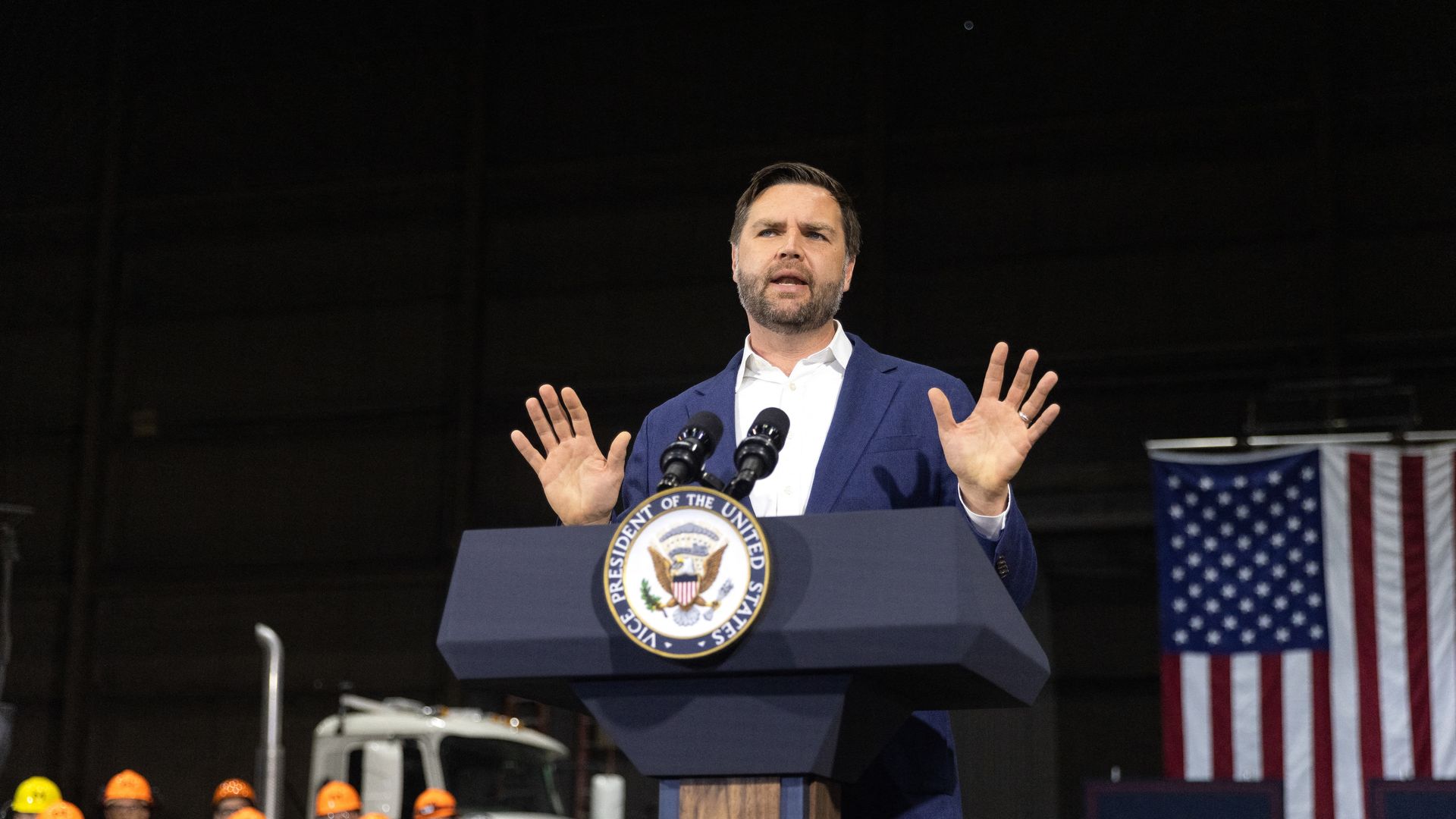 Man in blue suit speaking at a Vice President of the United States podium with dual microphones, raising hands, with workers in orange helmets and an American flag in the background.
