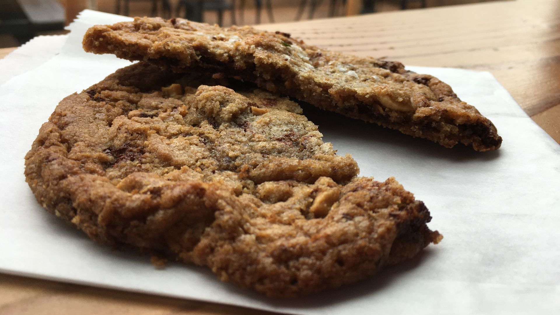 Two halves of a large chocolate chip cookie rest on a white paper bag.
