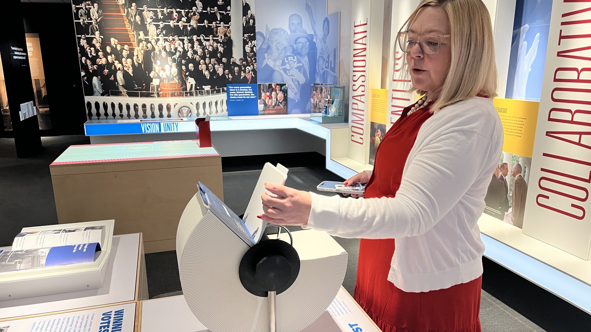 Woman in a red dress and white cardigan interacting with a rotating exhibit labeled "LBJ's Contact List" in a museum with large photos and words like "Compassionate" and "Collaborative."