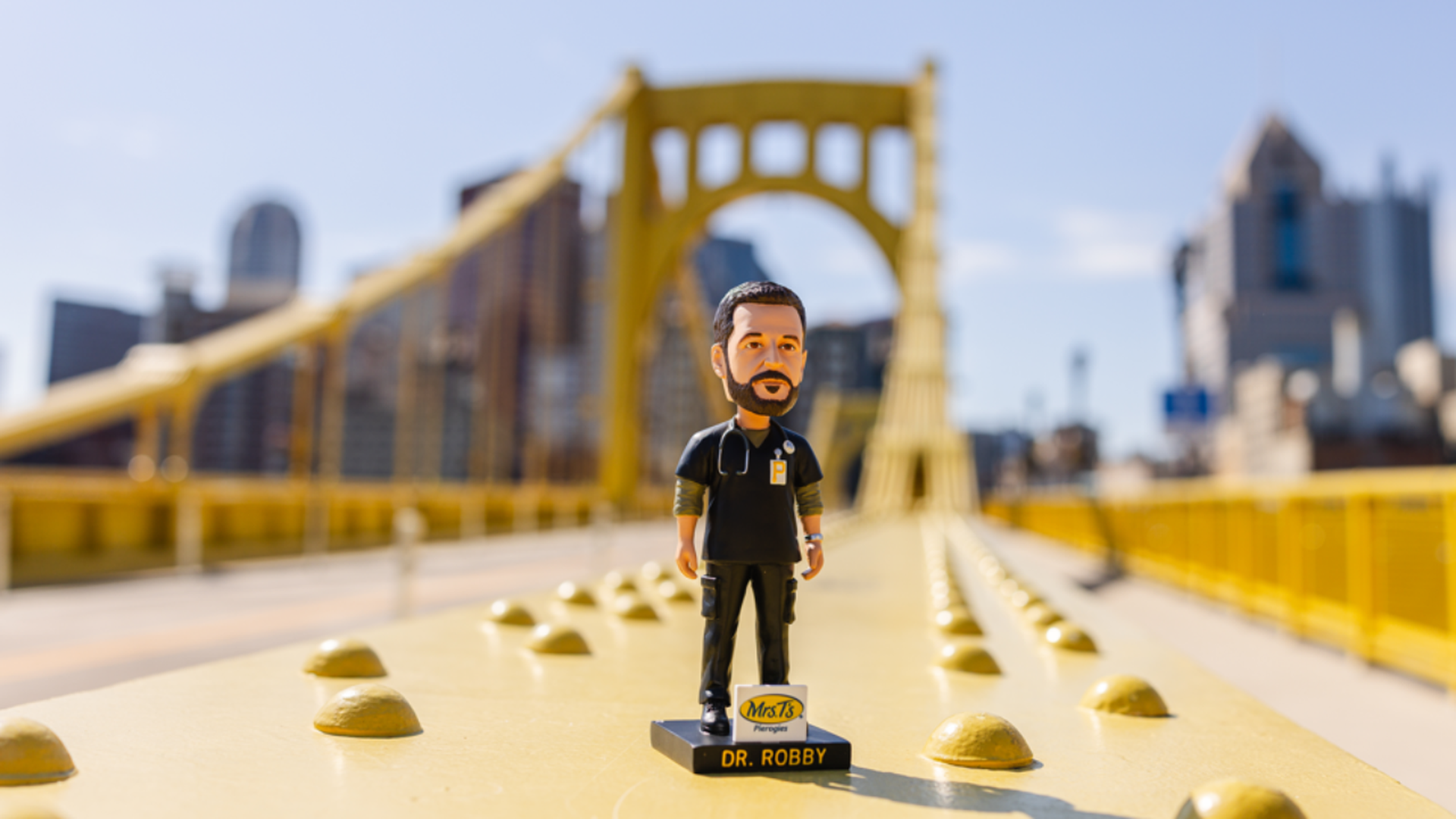 Bobblehead figure of Dr. Robby in black scrubs with a stethoscope, standing on a black base labeled DR. ROBBY on a bright yellow bridge, with a blurred city skyline and blue sky behind.