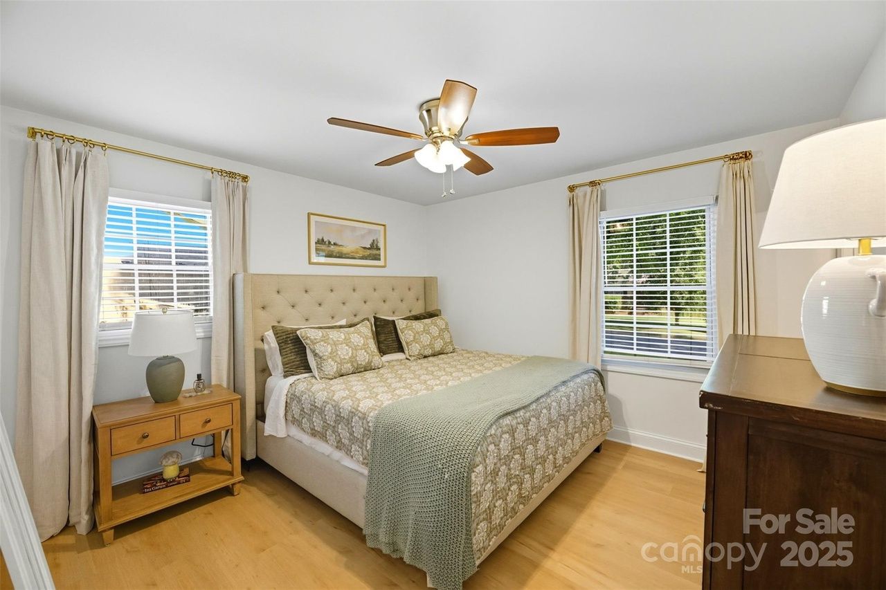 Bedroom with beige tufted headboard bed, patterned bedding, green throw, two windows with cream curtains, wooden nightstand with lamp, ceiling fan, wooden dresser, and landscape painting.