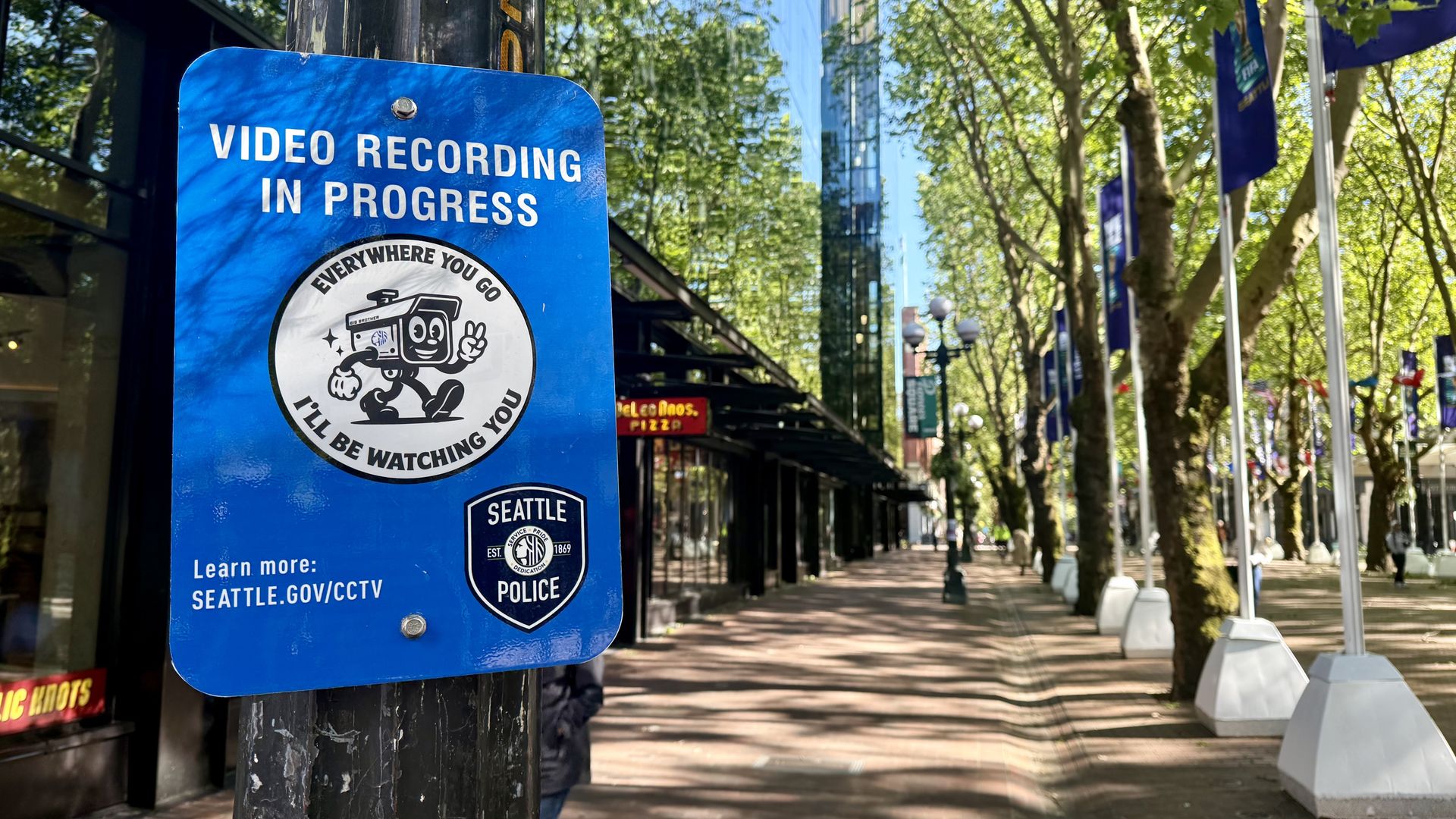 Blue sign on a worn black pole reads VIDEO RECORDING IN PROGRESS with a cartoon camera mascot; urban sidewalk with trees and purple banners in the background.