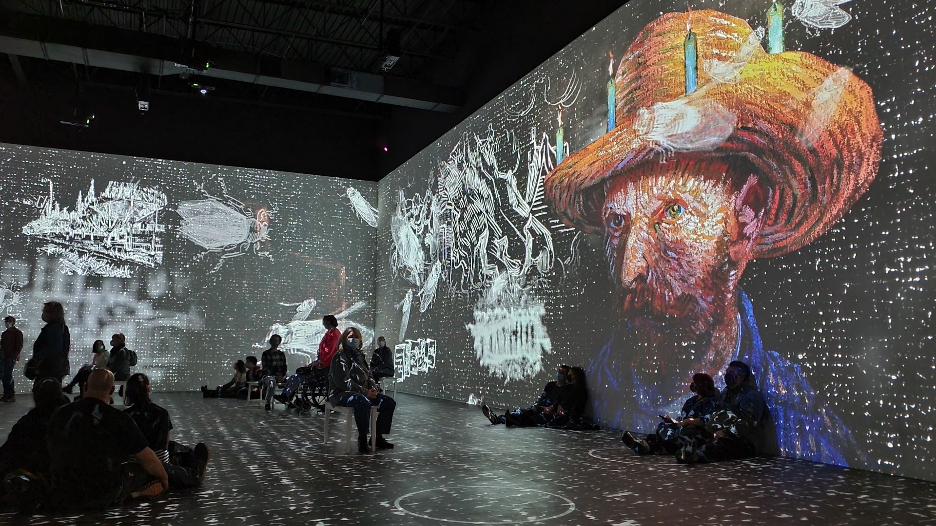 A portrait of Van Gogh is projected onto a wall while people sit on benches and the floor observing it