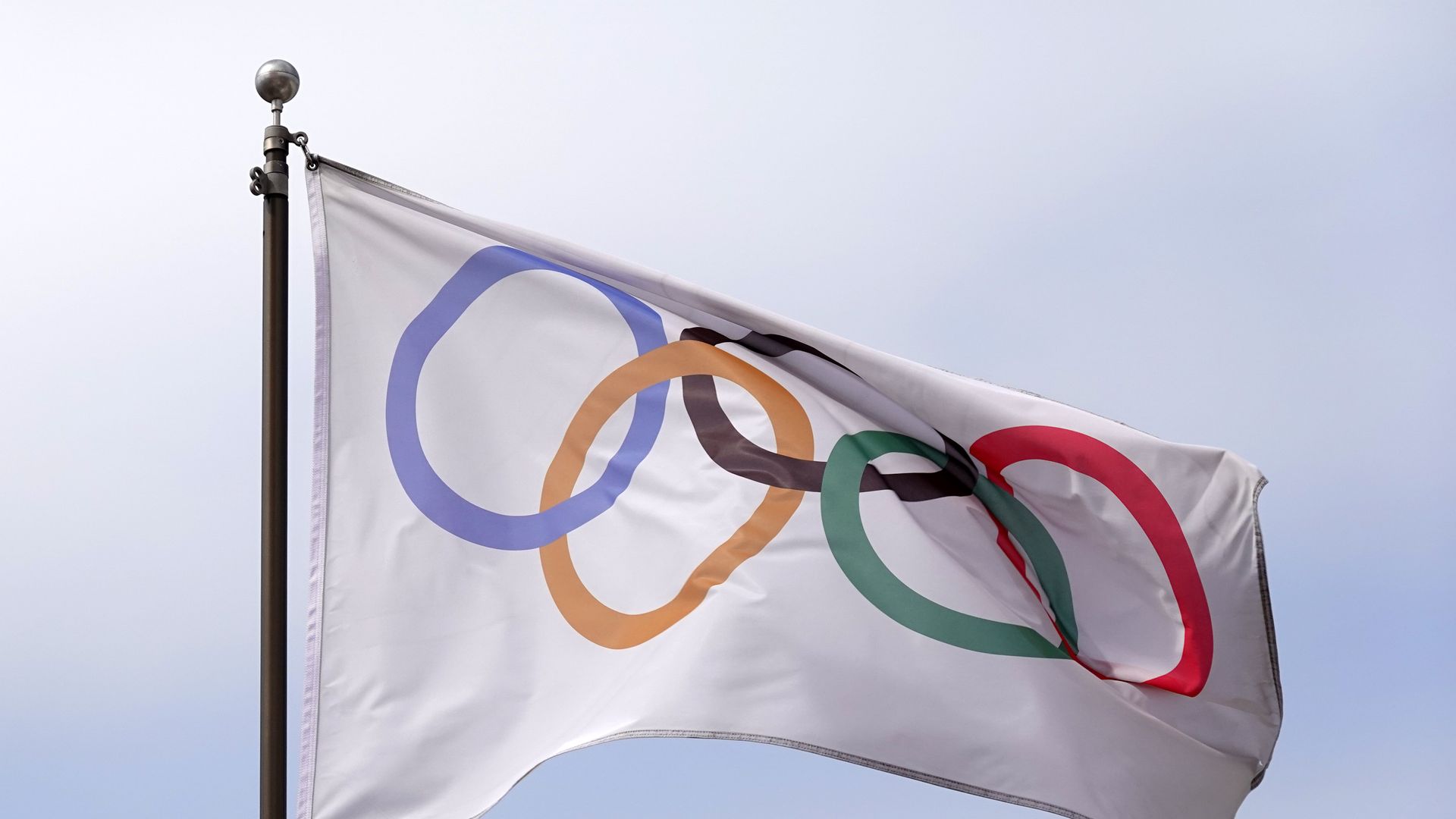 White flag with five interlocking Olympic rings in blue, yellow, black, green, and red, flying on a flagpole against a clear sky.