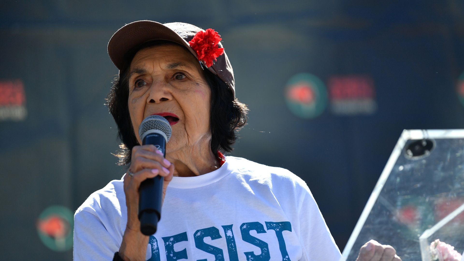 Dolores Huerta wears a ballcap with a red flower while holding a microphone next to a clear podium. Her white T shirt says "resist" in blue capital letters.