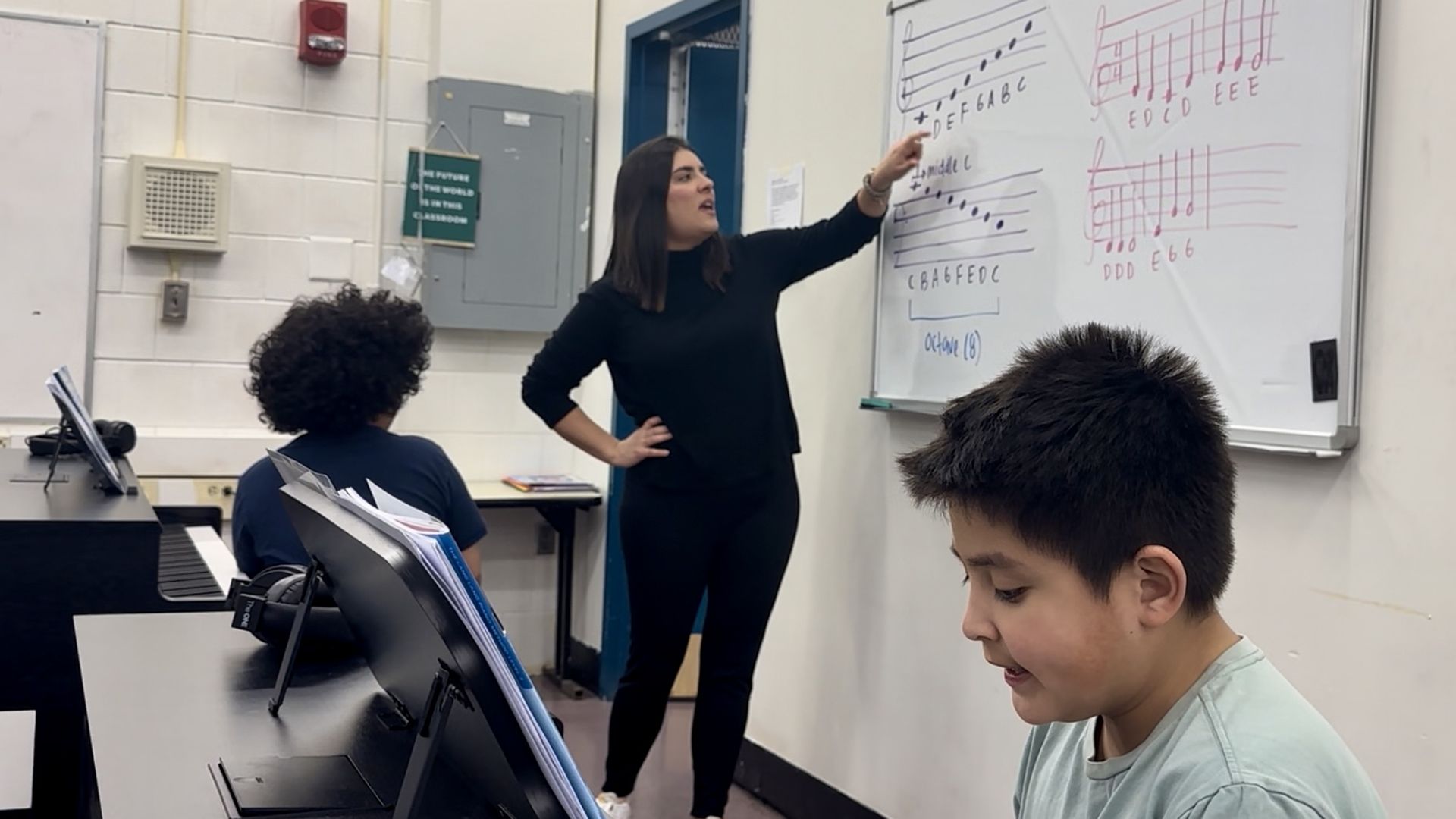 Music teacher Virginia Lopez Alvarez, center, points to a whiteboard showing a "C" scale, as one student, at a piano, left, looks on, and a second student, right, looks at his piano keys.