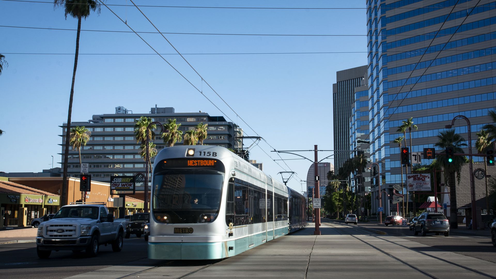 A silver light rail train with teal trimming along the bottom is next to cars stopped at a traffic signal with tall buildings along the street behind it. 