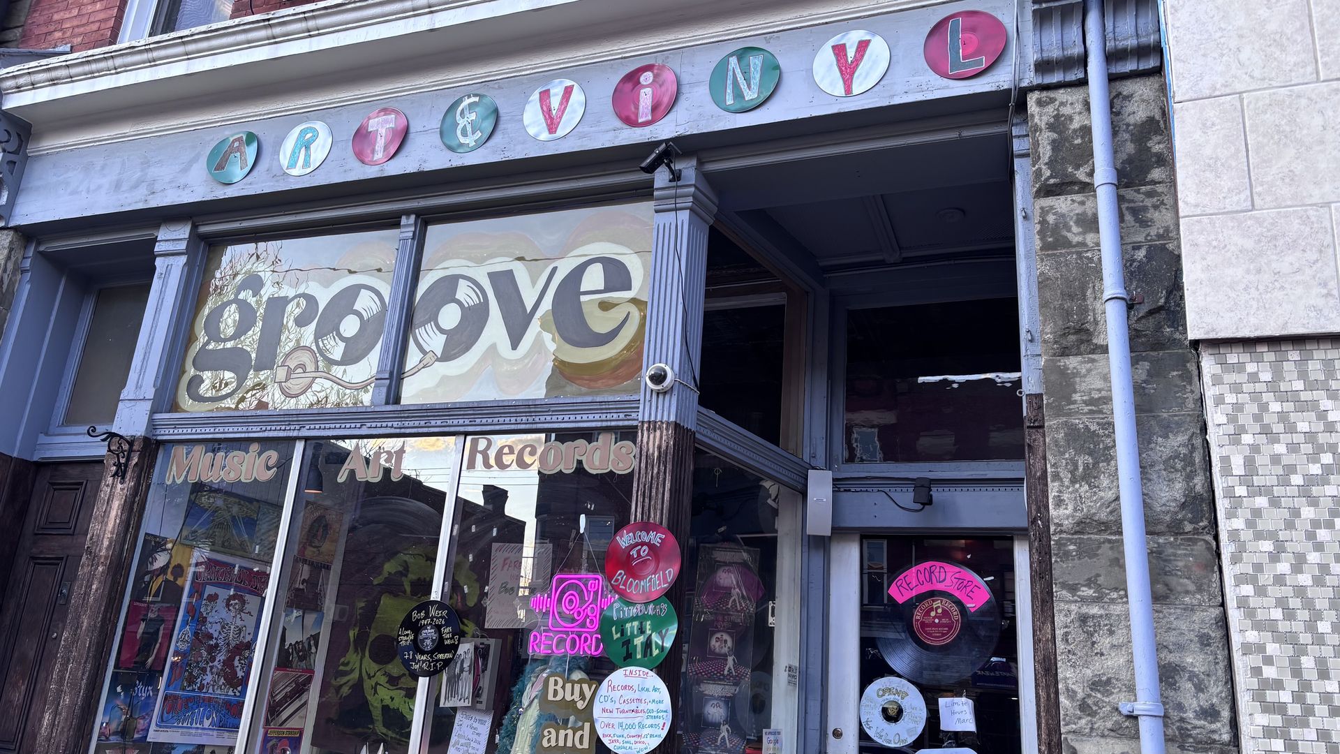 Front of a vintage store with round colorful signs spelling ART & VINYL, large 'groove' text on window. Posters and record-themed decals decorate the glass storefront.
