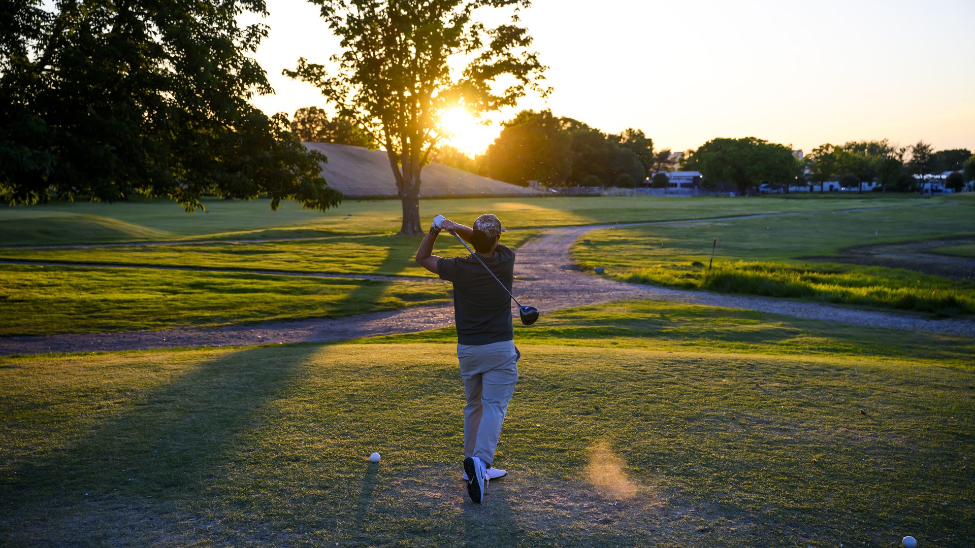 A golfer in a dark shirt and light shorts swings a club on a sunlit golf course at sunset, with a bright sun behind trees, a dirt cart path, and a ball near the tee.