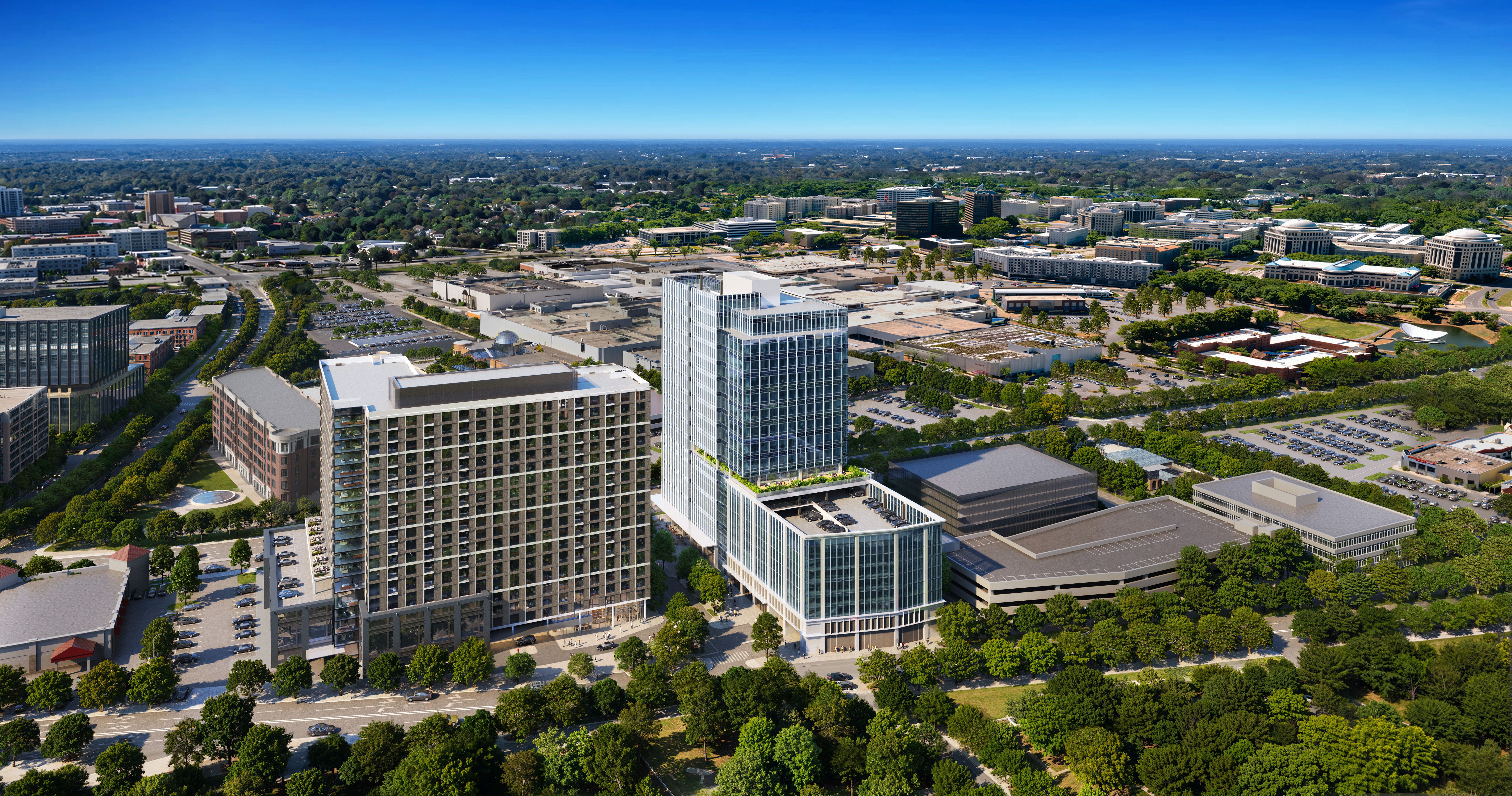 Panoramic aerial view of a modern city with a tall glass skyscraper in the foreground, surrounded by mid-rise buildings, tree-lined streets, parking lots, and a clear blue sky.