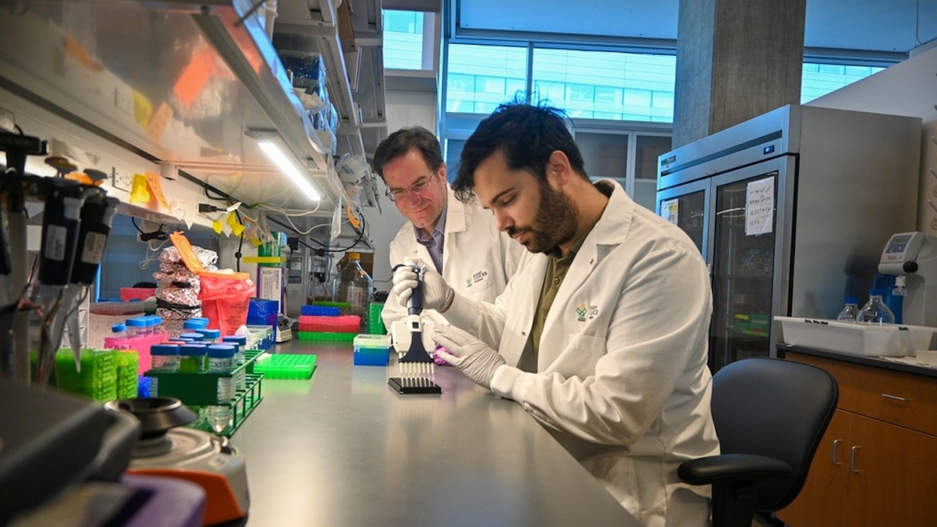 Two people are picture in a photo where they sit in a medical lab and using instruments for a blood test.