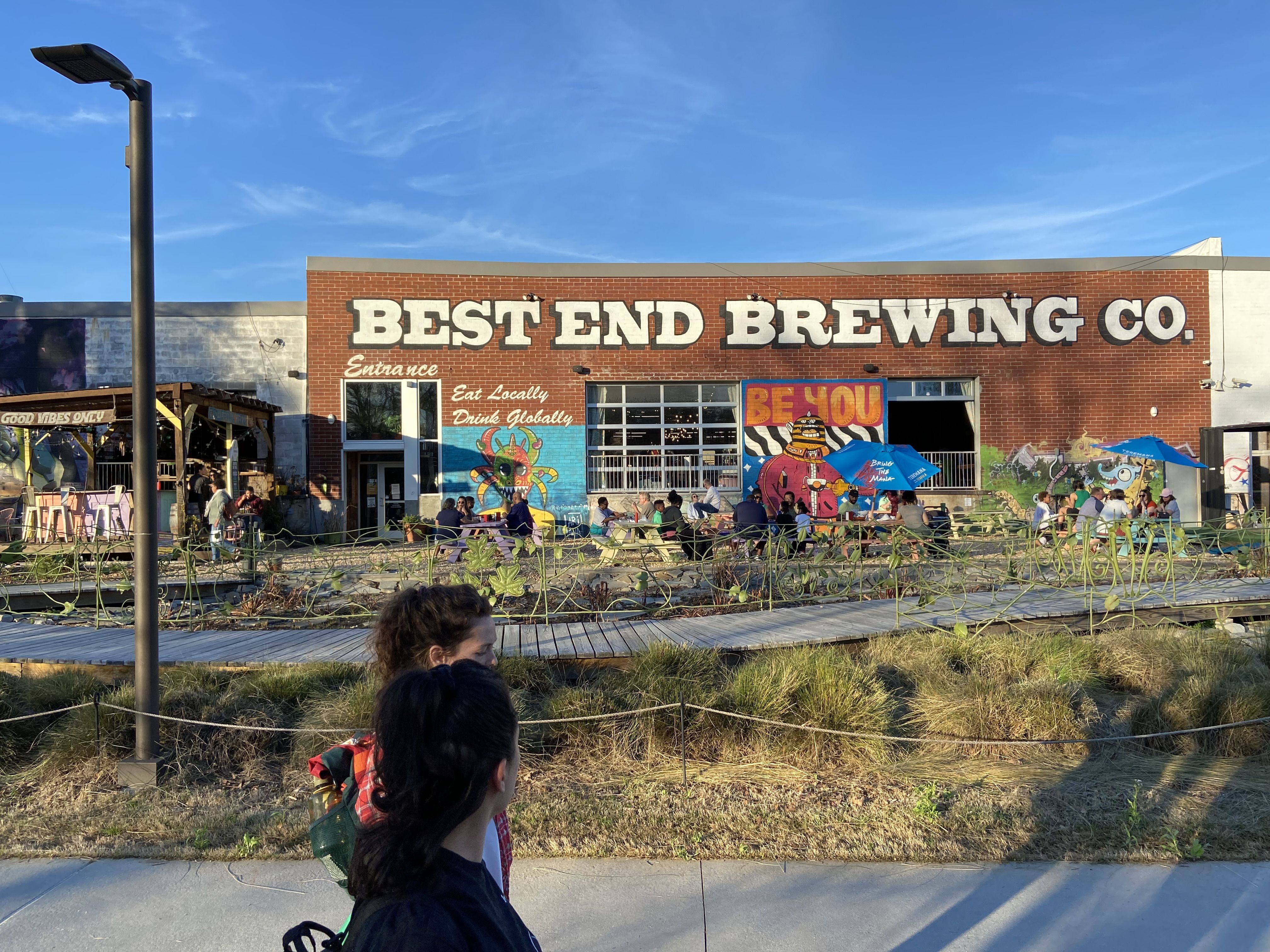 Two people walk on a paved bike and pedestrian trail in front of a building with "BEST END BREWING" in capital letters
