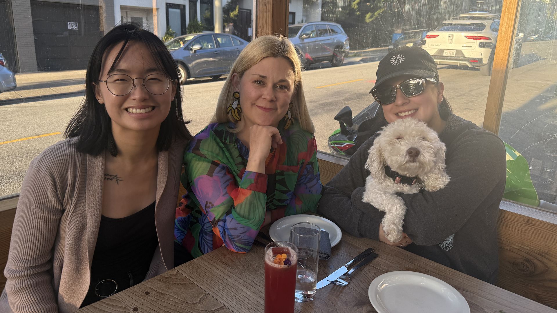 Three friends sit at a wooden outdoor table under a glass shelter. A blonde in a colorful shirt sits middle; to the right, a cap-wearing woman holds a fluffy white dog. Cars are on the street behind.