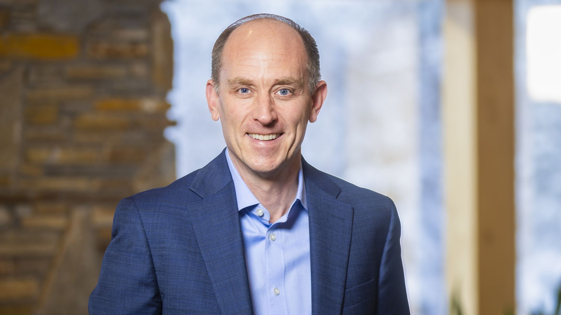 Smiling bald man with blue eyes wearing a blue suit and light blue shirt stands indoors with blurred windows and stone wall in the background.