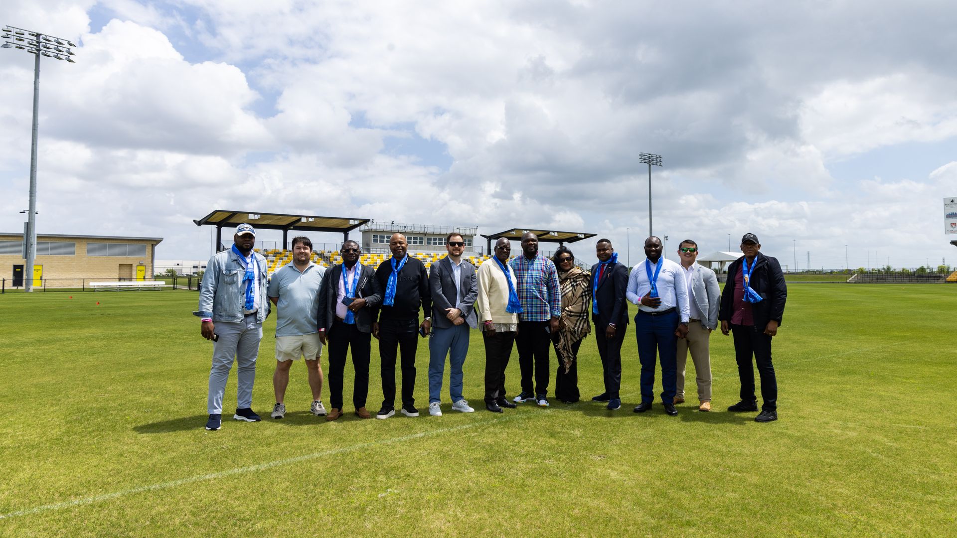 Group of adults standing shoulder-to-shoulder on a green sports field, wearing blue scarves. Stadium seating and tall floodlights rise behind them under a cloudy blue sky.