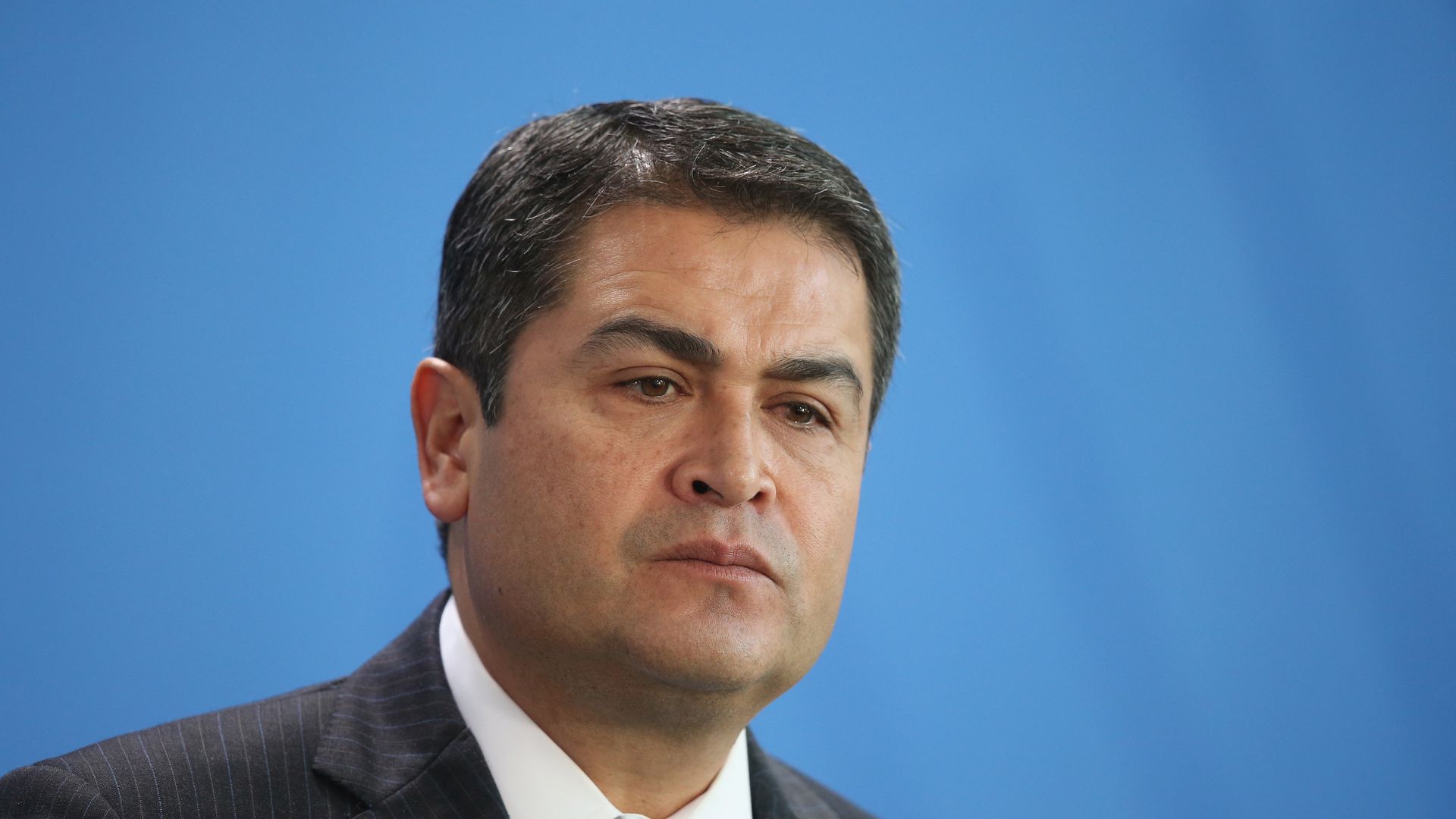 Juan Orlando Hernandez looks off to the side. the background behind him is sky blue.