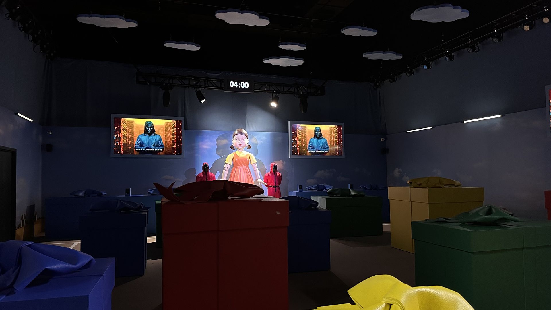 A photo of a large doll at the back of a room and several large fake presents closer to the front, like in Squid Game