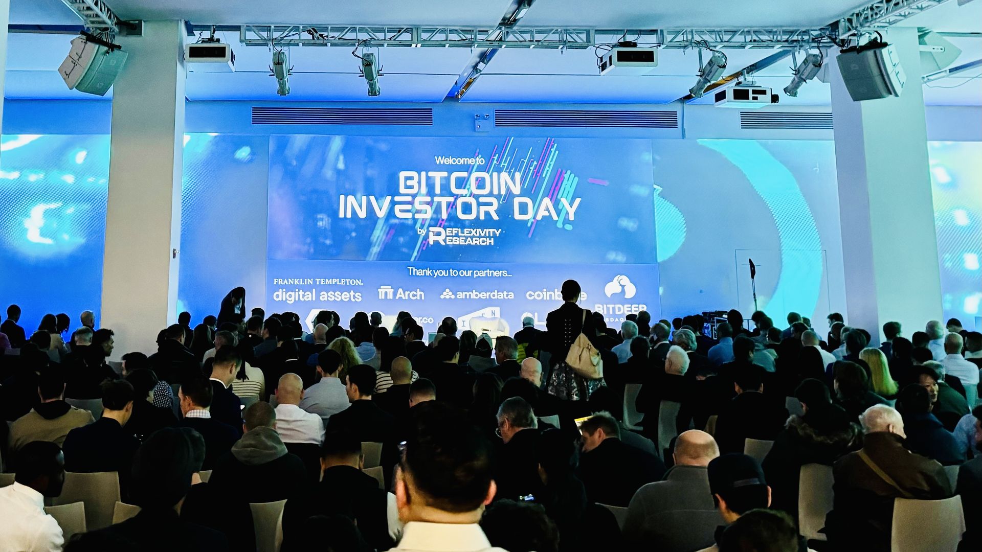 A picture from the back of a room toward a stage with a sign reading Bitcoin Investor Day
