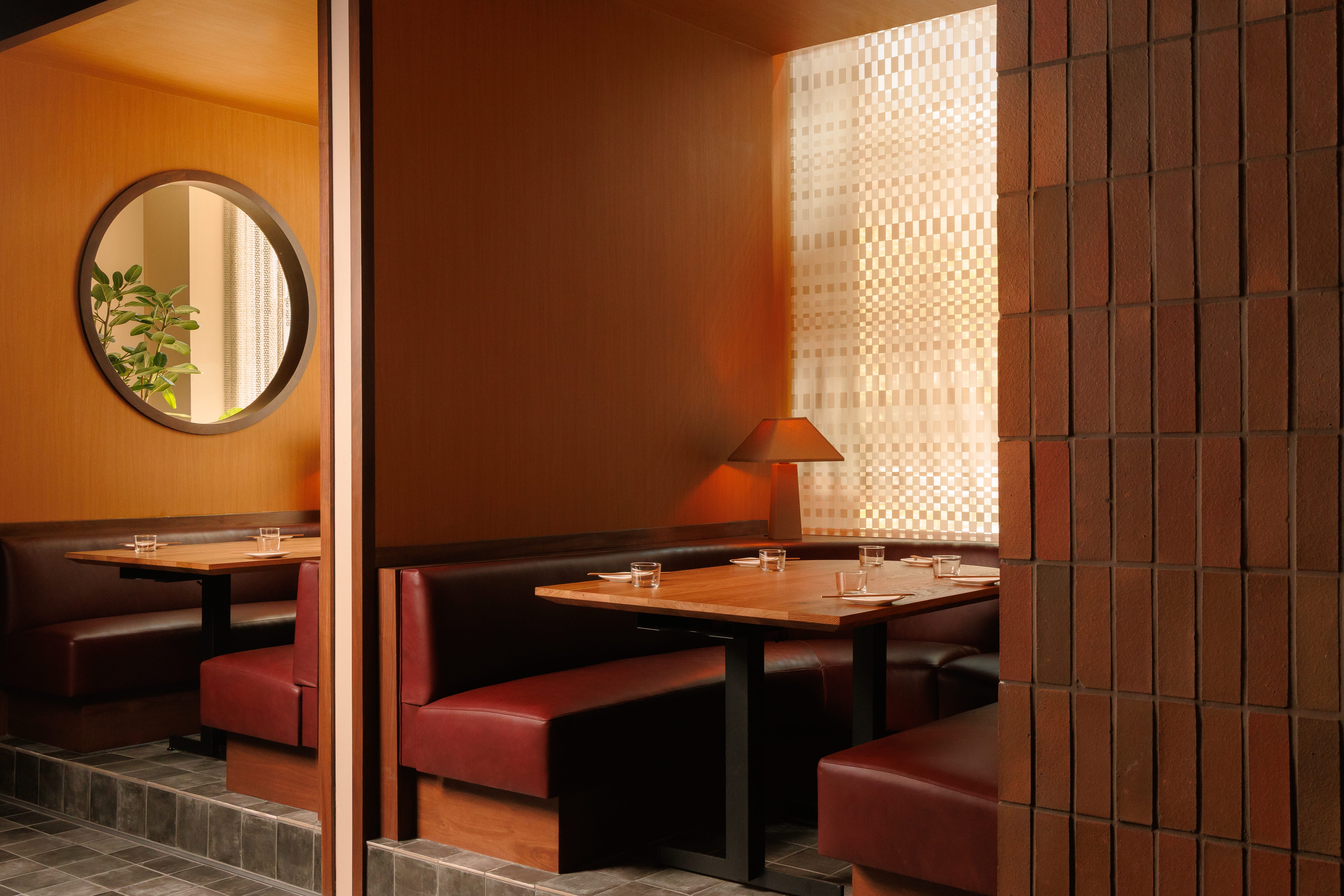Warm-lit restaurant interior with wooden walls and brown brick columns, red leather booths, a round mirror reflecting a green plant, and a light, checkered wall near the seating.