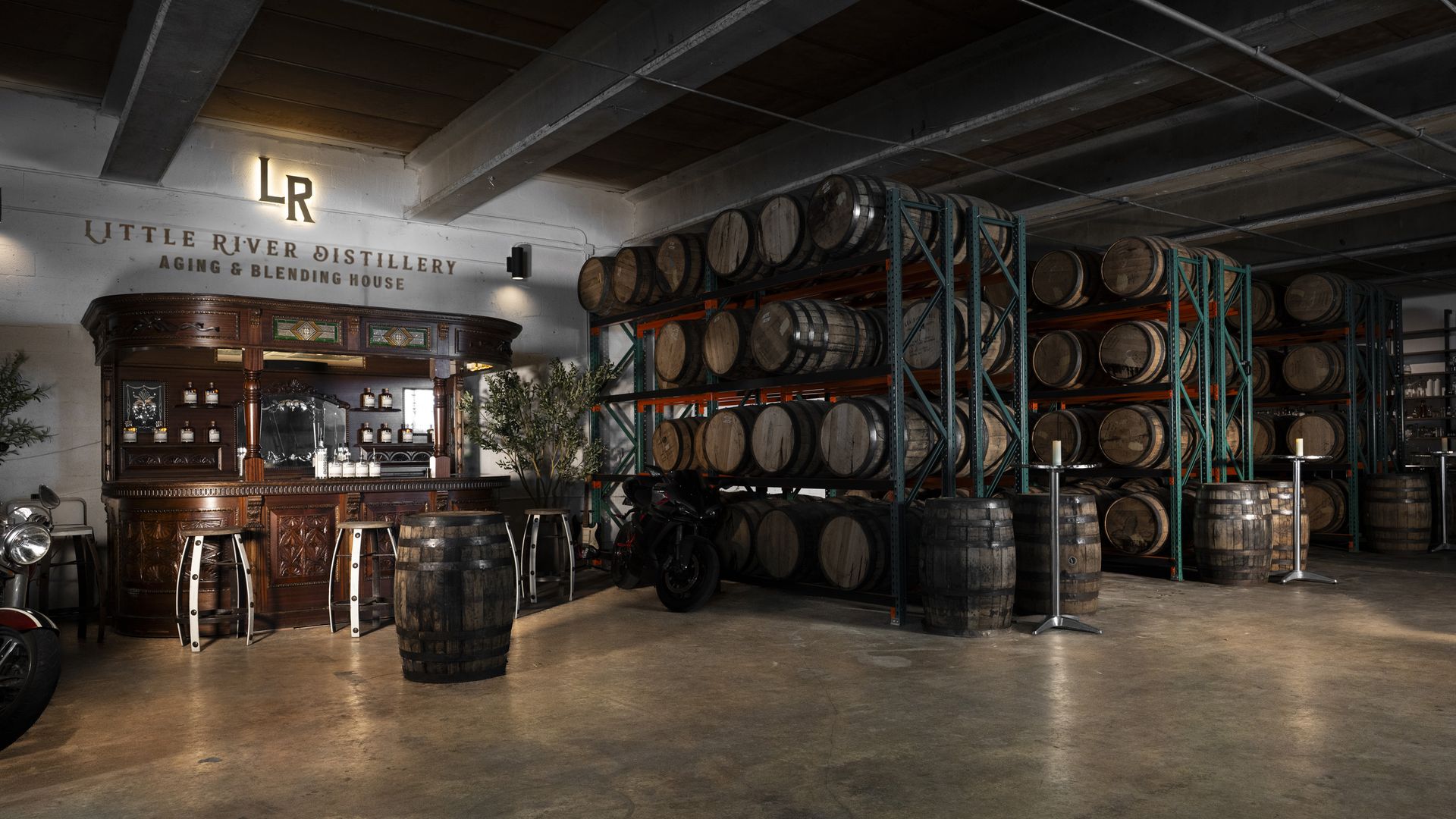Industrial distillery interior with rows of wooden barrels on green metal racks, an ornate wooden bar to the left, and a wall sign reading \"Little River Distillery\" with \"Aging & Blending House\" beneath.