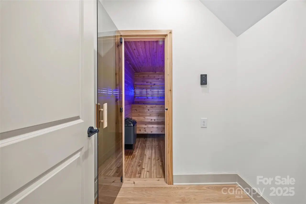 View through an open door into a wooden sauna room with blue LED lighting and a heater, adjacent to a plain white wall with a thermostat and outlet.