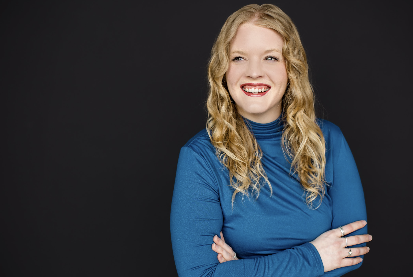 Portrait of a smiling blonde woman with long, wavy hair wearing a blue turtleneck, arms crossed, against a dark backdrop.