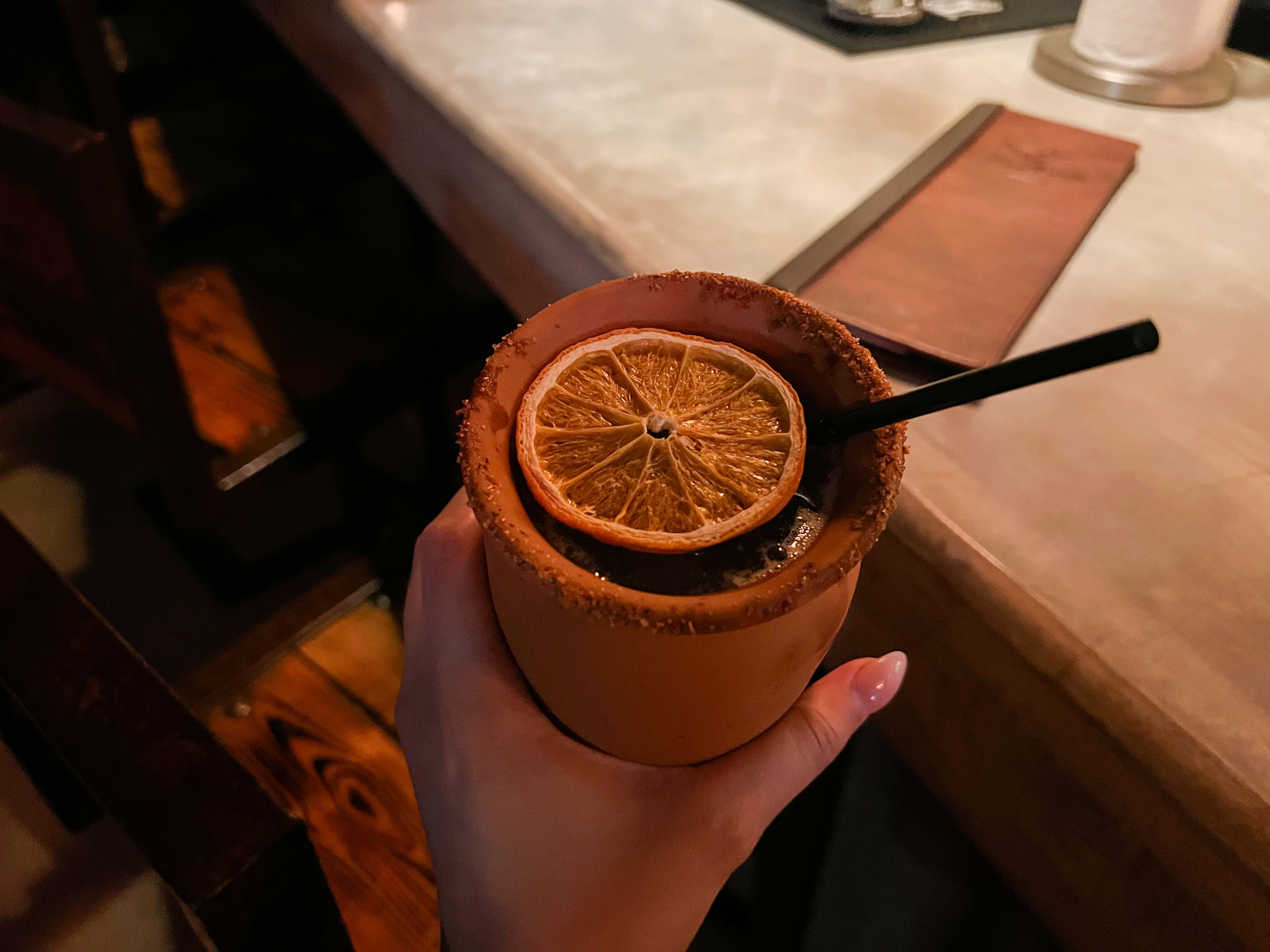 A hand holding a cocktail served in a teracota cup, garnished with an orange slice.