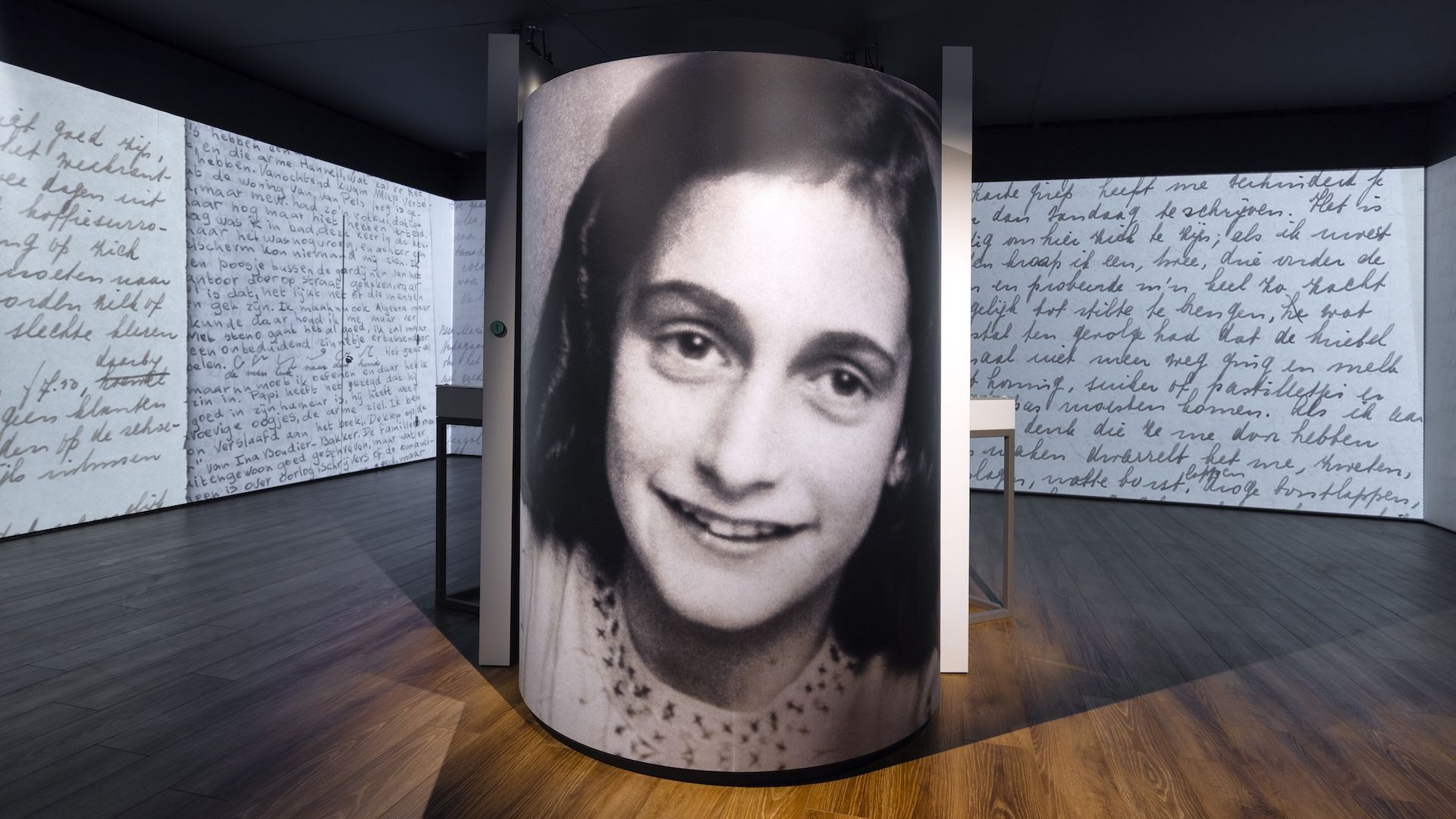 Dim museum gallery with a tall cylindrical column printed with a large black-and-white portrait of a smiling girl. Surrounding panels show cursive handwriting in multiple languages; wooden floor.
