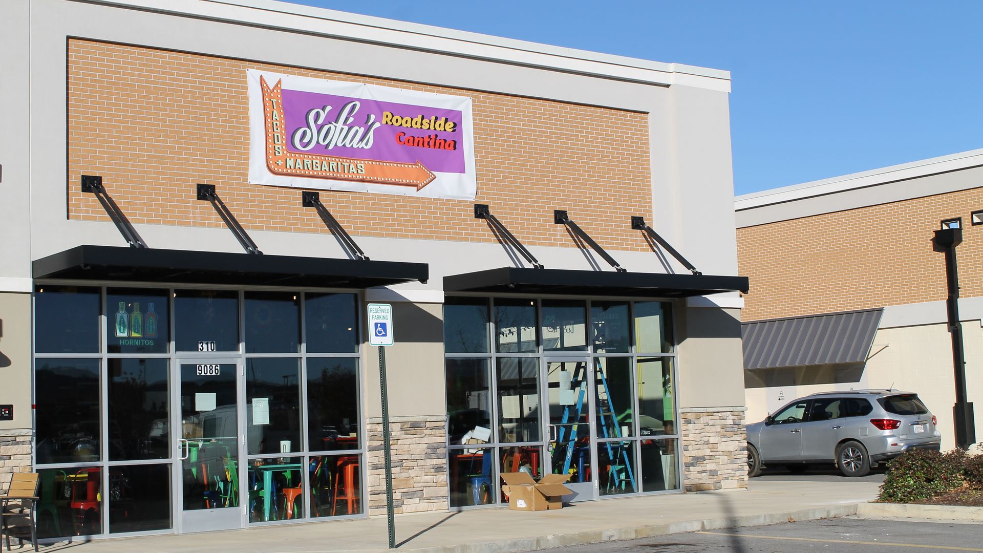 Exterior of Sofia's Roadside Cantina with a sign featuring an orange arrow on purple background, large glass doors, colorful chairs inside, a reserved parking sign, and a silver SUV parked nearby.