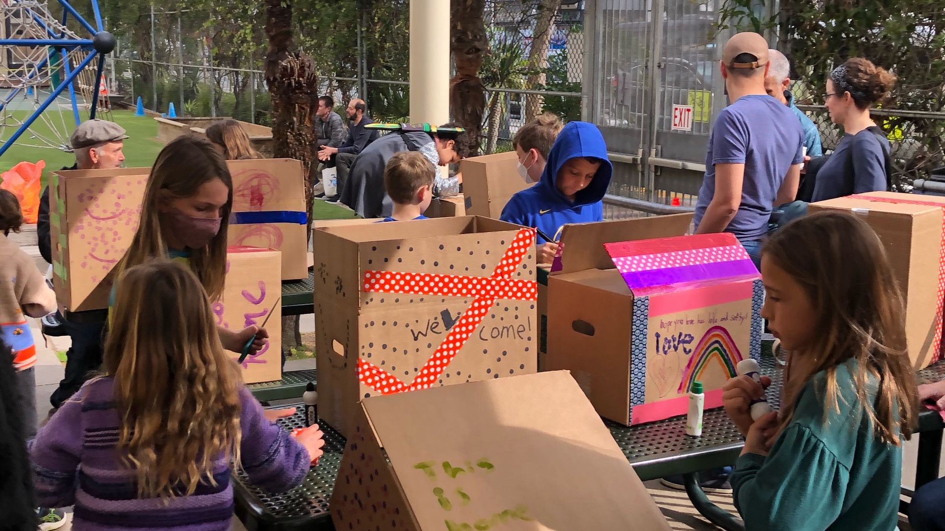 Children sit at picnic tables decorating cardboard boxes on a school playground.