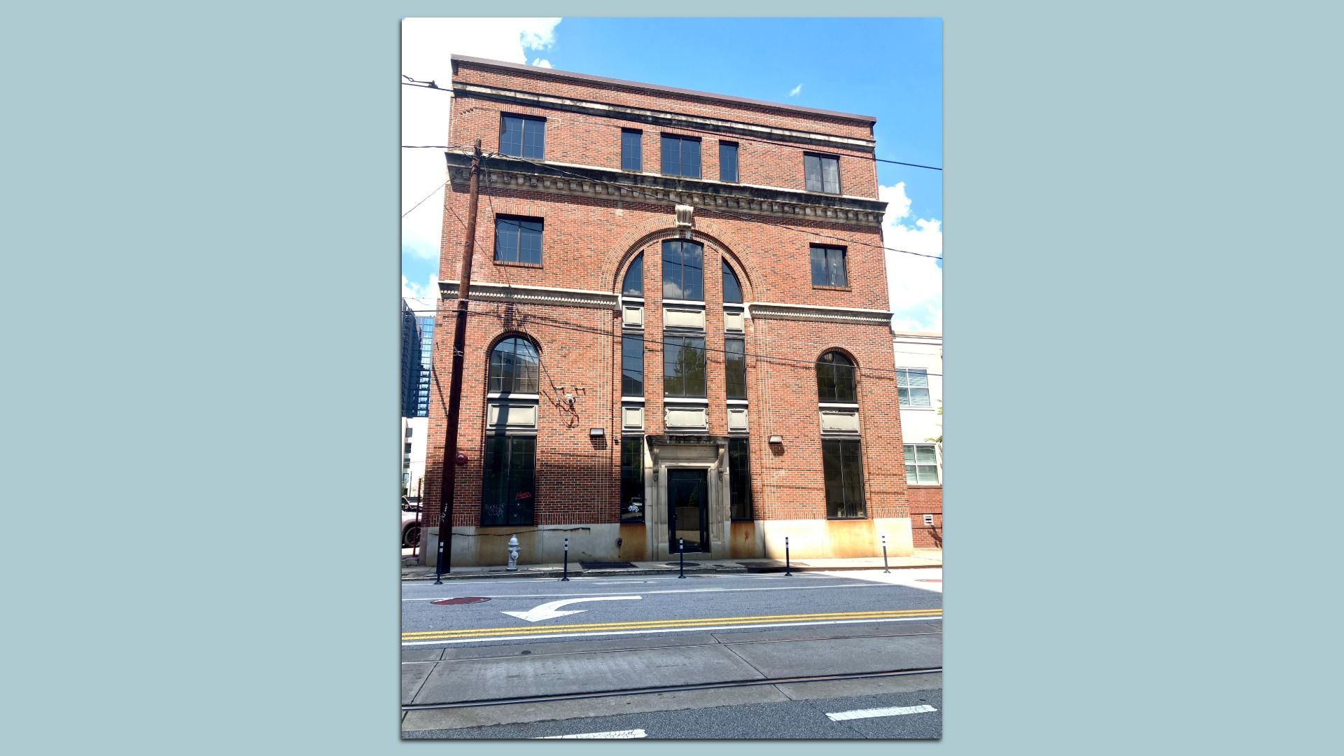 Four-story red brick building with large black-framed windows, stone accents, and arched windows on the second floor, situated on a street with tram tracks and a left turn arrow.