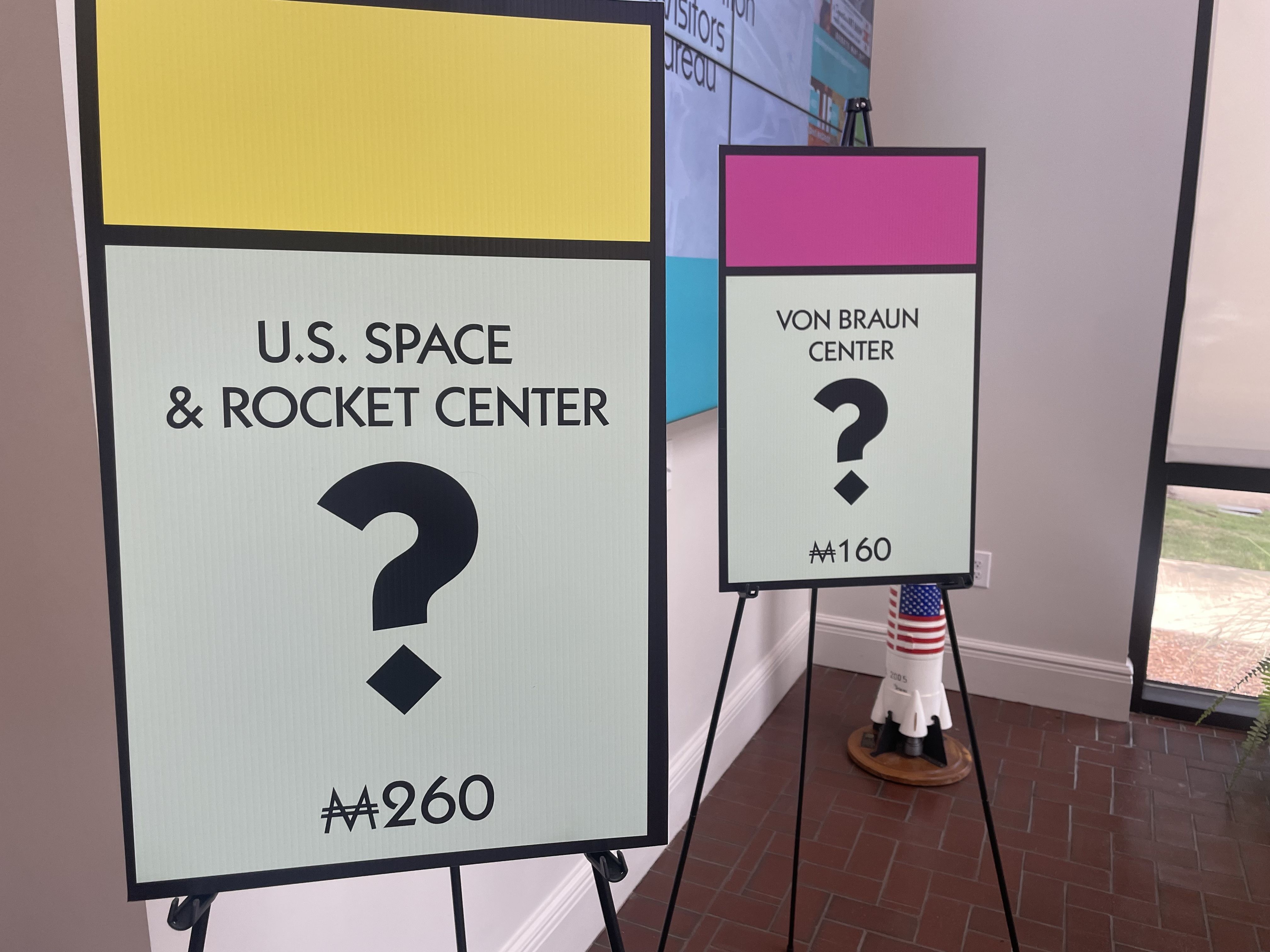 Indoor exhibit with sign boards in bold color blocks. Left board reads "U.S. SPACE & ROCKET CENTER" under a yellow panel; right board reads "VON BRAUN CENTER" with a pink top and a large question mark.