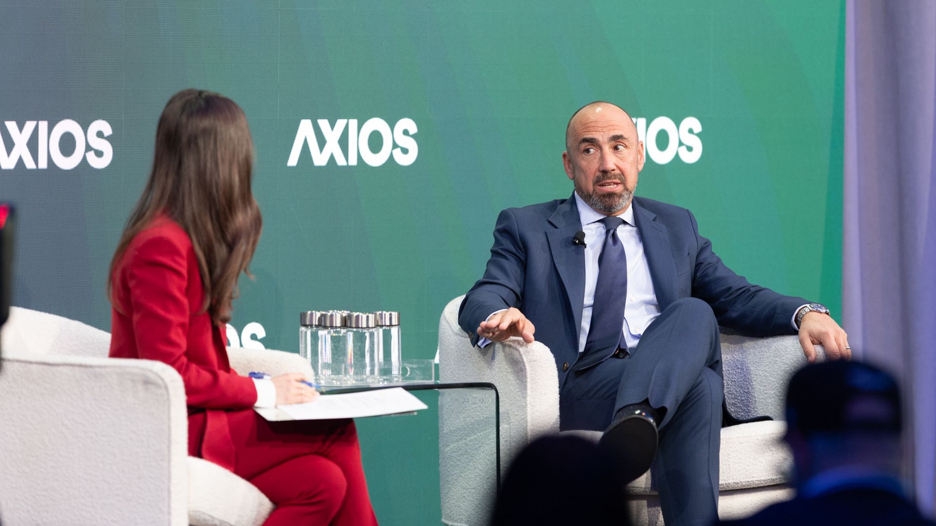 Panel discussion on a stage with a green Axios backdrop. A man in a navy suit speaks, gesturing with his hand; a woman in red sits opposite with notes and a glass table between them.