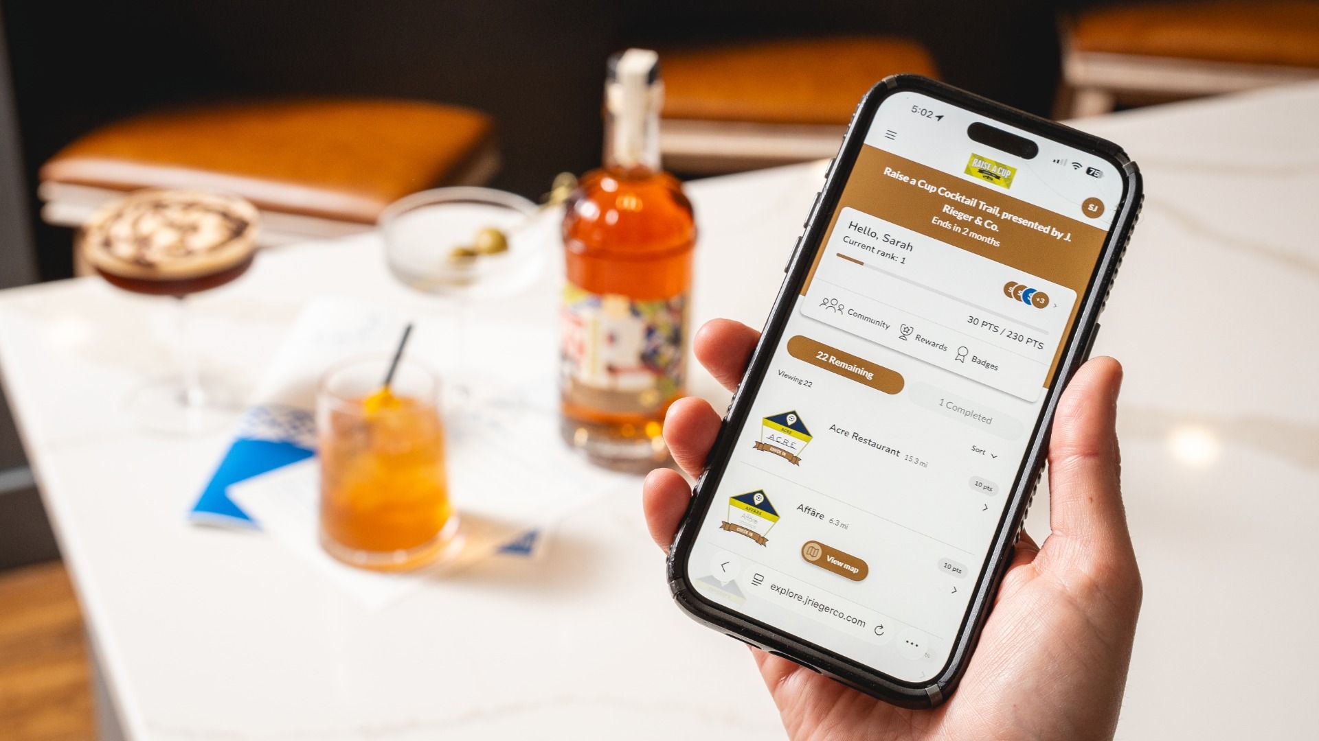 A hand holds a smartphone showing a brown-themed app screen with the title "Raise a Cup Cocktail Trail"; on a white table there is an amber liquor bottle and a cocktail, with a blurred background.