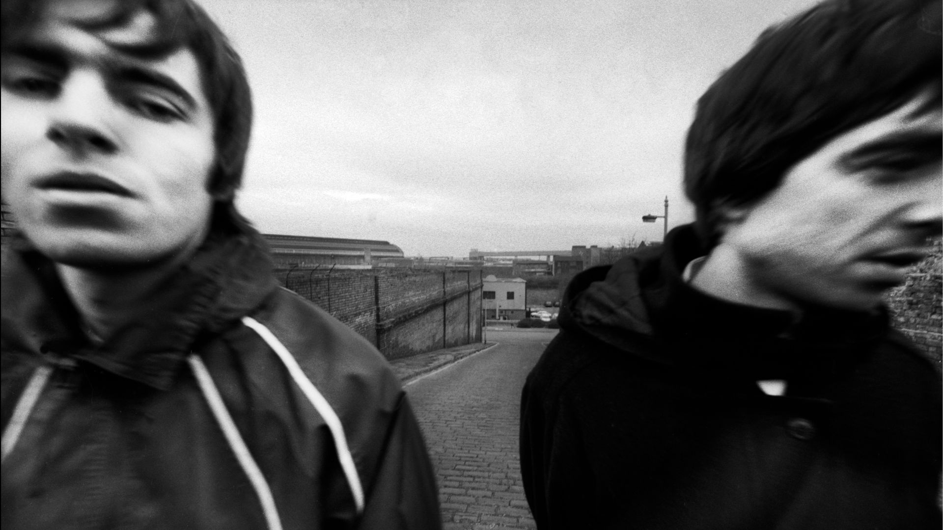 Black-and-white close-up of two people in jackets, faces blurred, standing on a brick road with a fence and industrial buildings visible in the background under a gray sky.