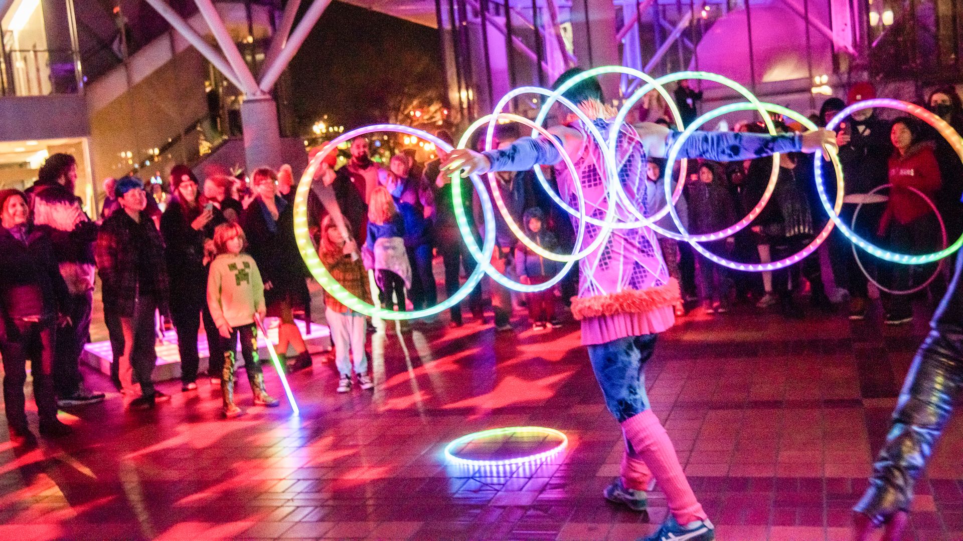 A crowd at night watches a woman spinning illuminated hoops.