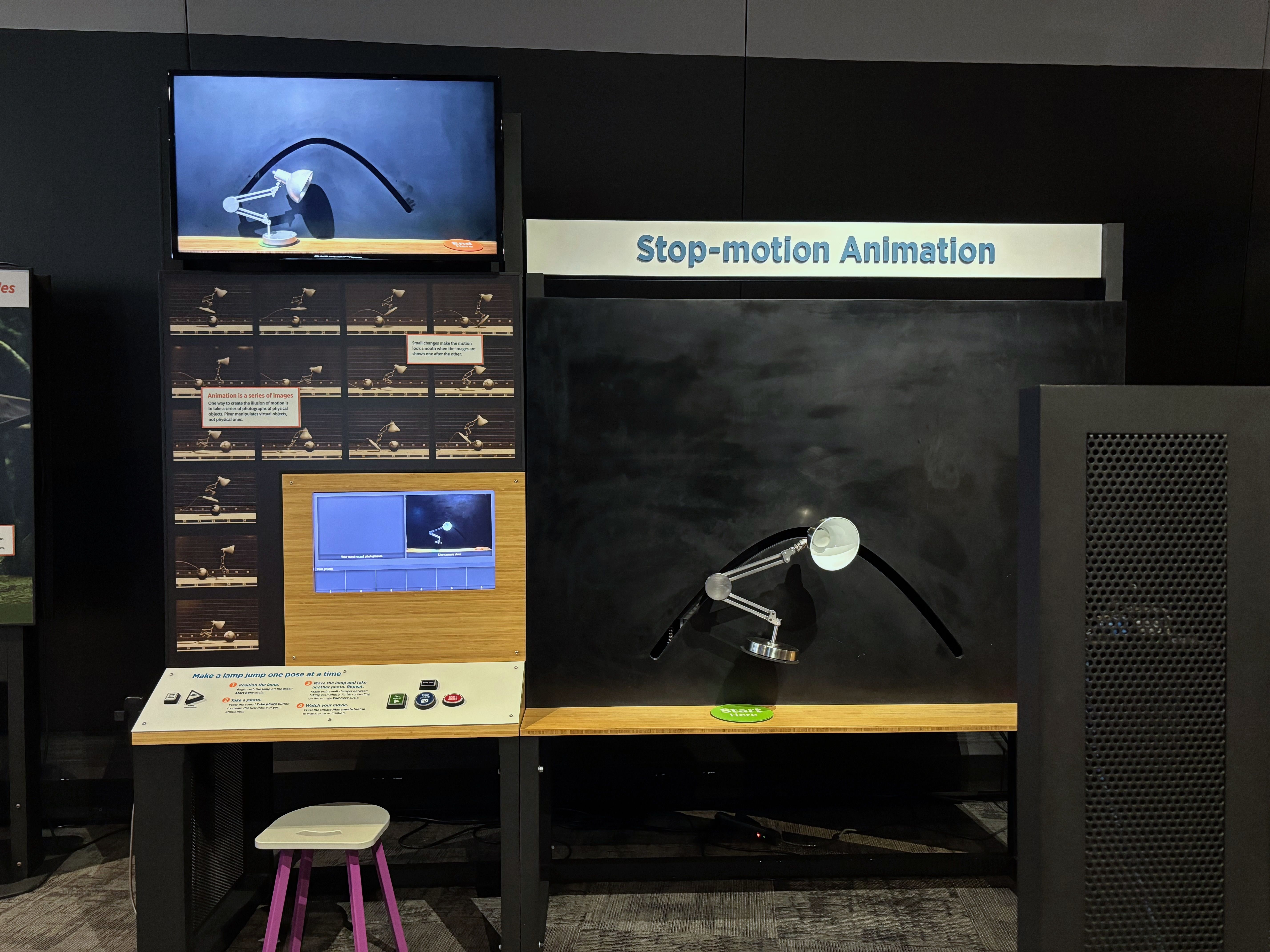 Interactive stop-motion animation exhibit with a white desk lamp on a black curved track, screens showing animation frames, and instructions for making a lamp jump pose by pose.