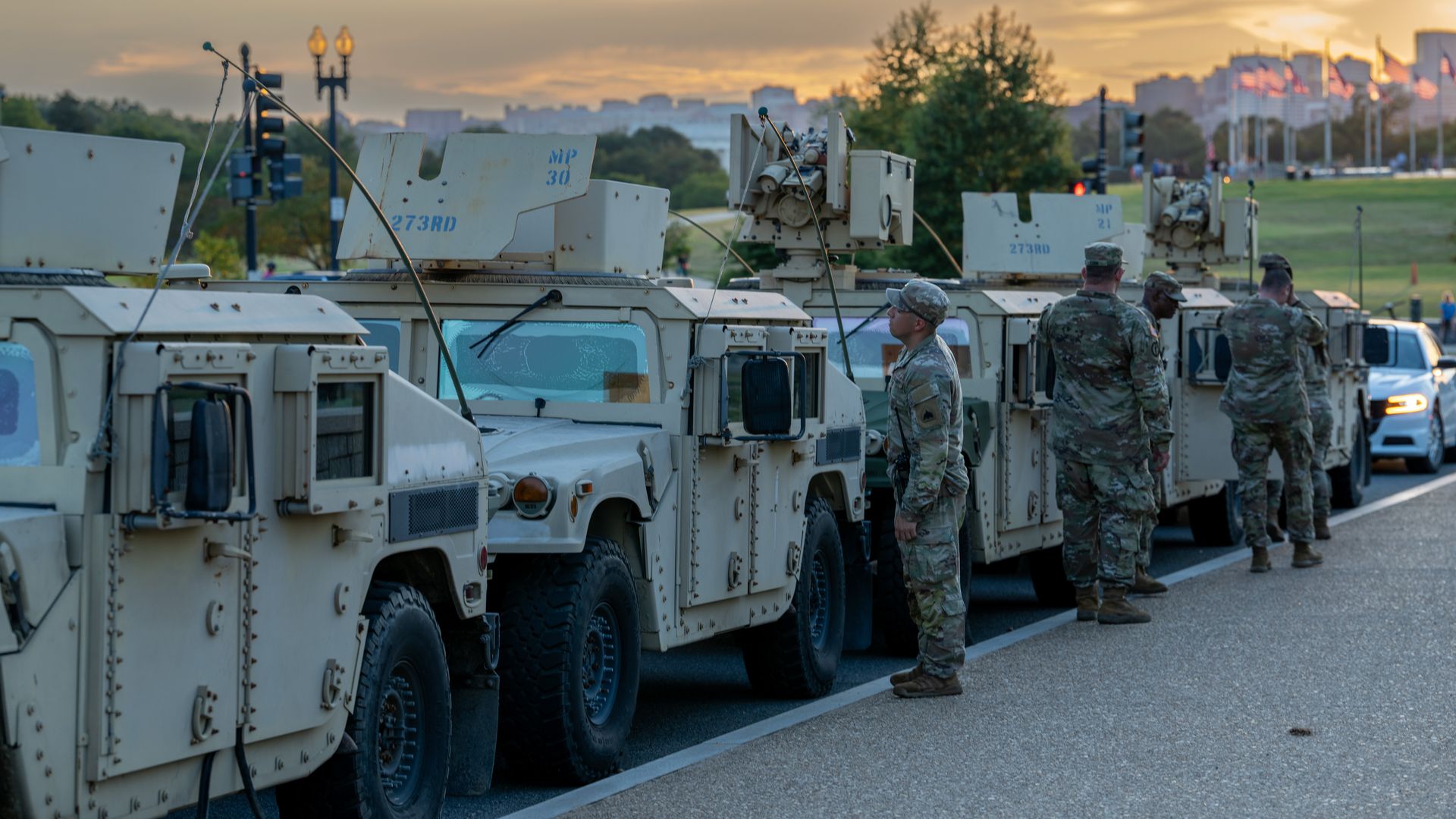 Four tan military Humvees lined up on a road at sunset with five soldiers in camouflage uniforms standing beside them and American flags in the background.