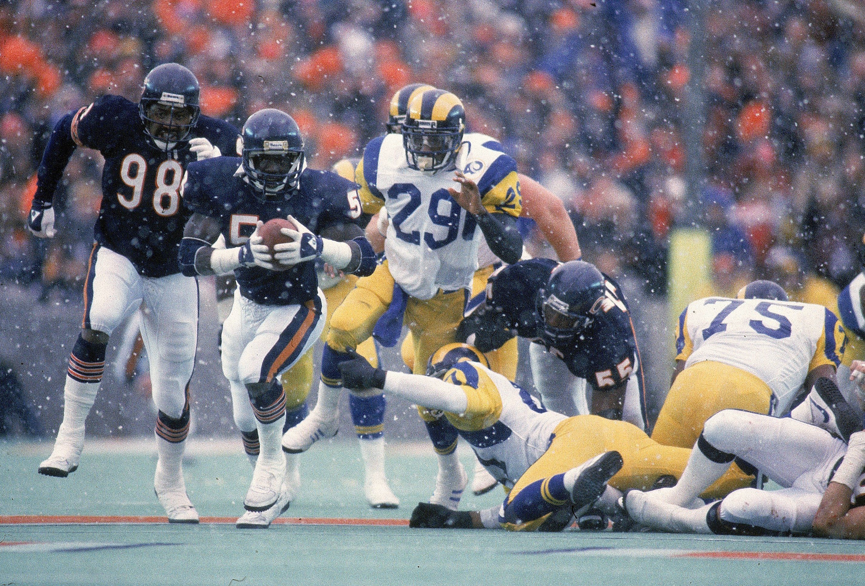 Snowy football game with players in navy and orange and others in white and yellow uniforms, a player in navy catching the ball mid-action with others running and on the ground.